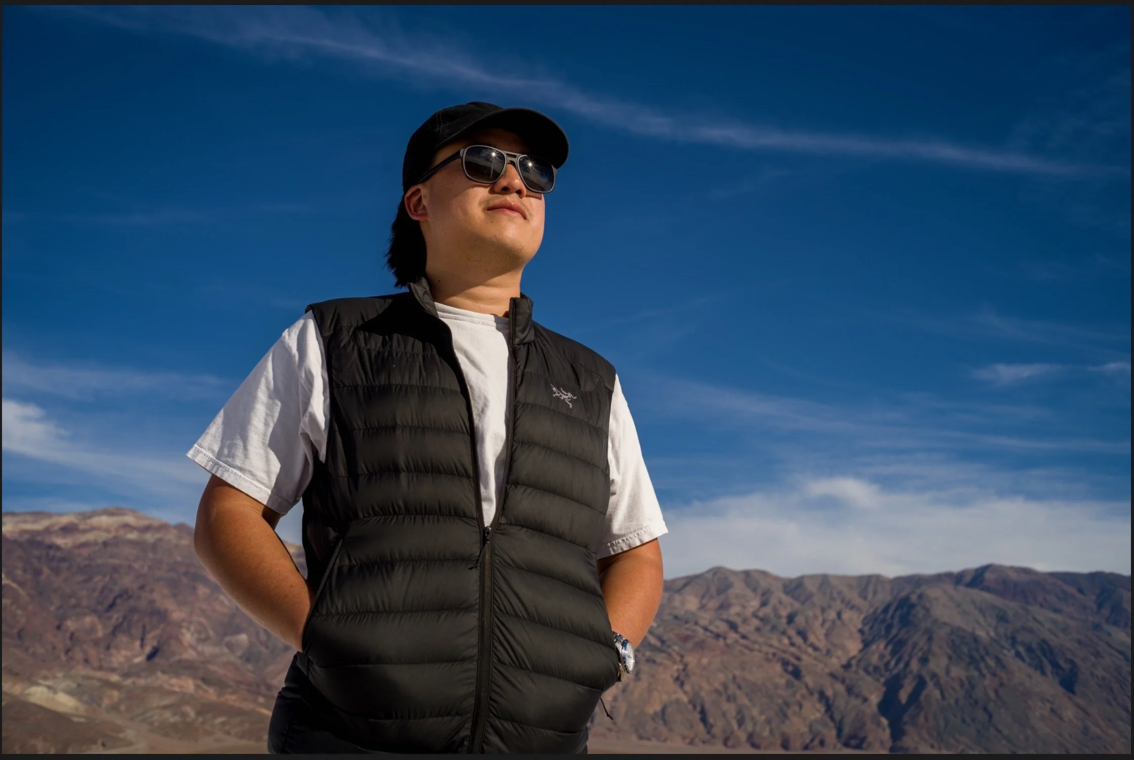 A man in sunglasses and a black cap standing outdoors in front of mountains under a blue sky.