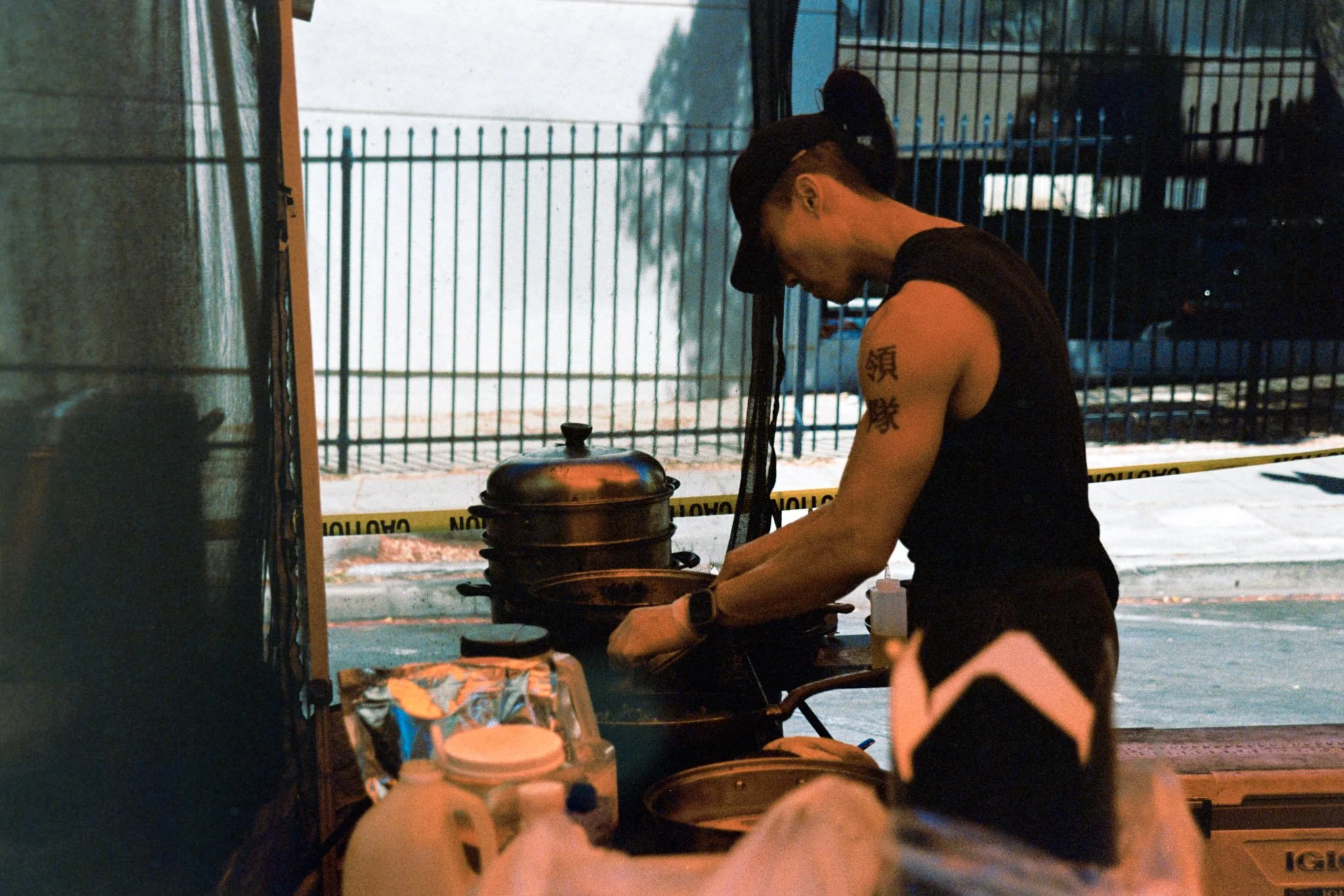 A person with tattoos on their arm preparing food at an outdoor counter, with a black fence and trees in the background and caution tape on the ground.