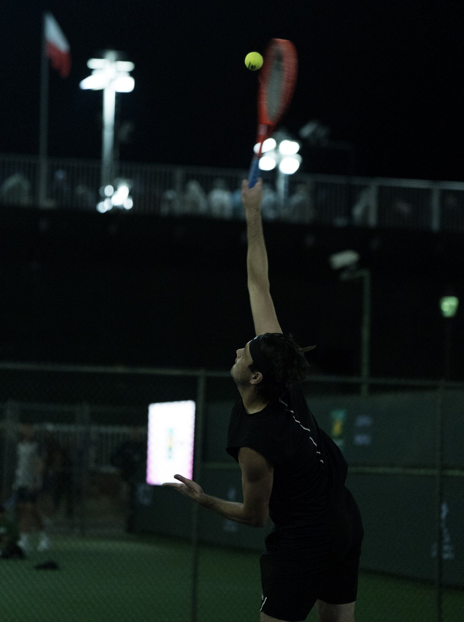 A person playing tennis at night, reaching up to hit a yellow tennis ball with a red racquet, with tennis courts and bright lights in the background.