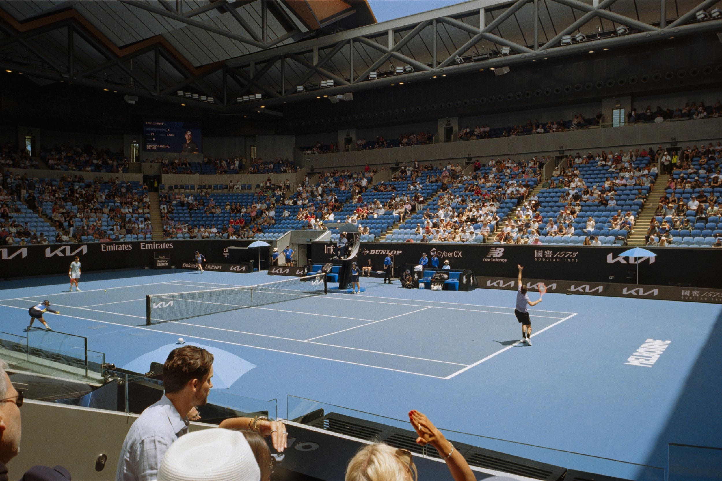 Spectators watching a tennis match at a stadium with blue court, with some players in action and advertisements on the sidelines.