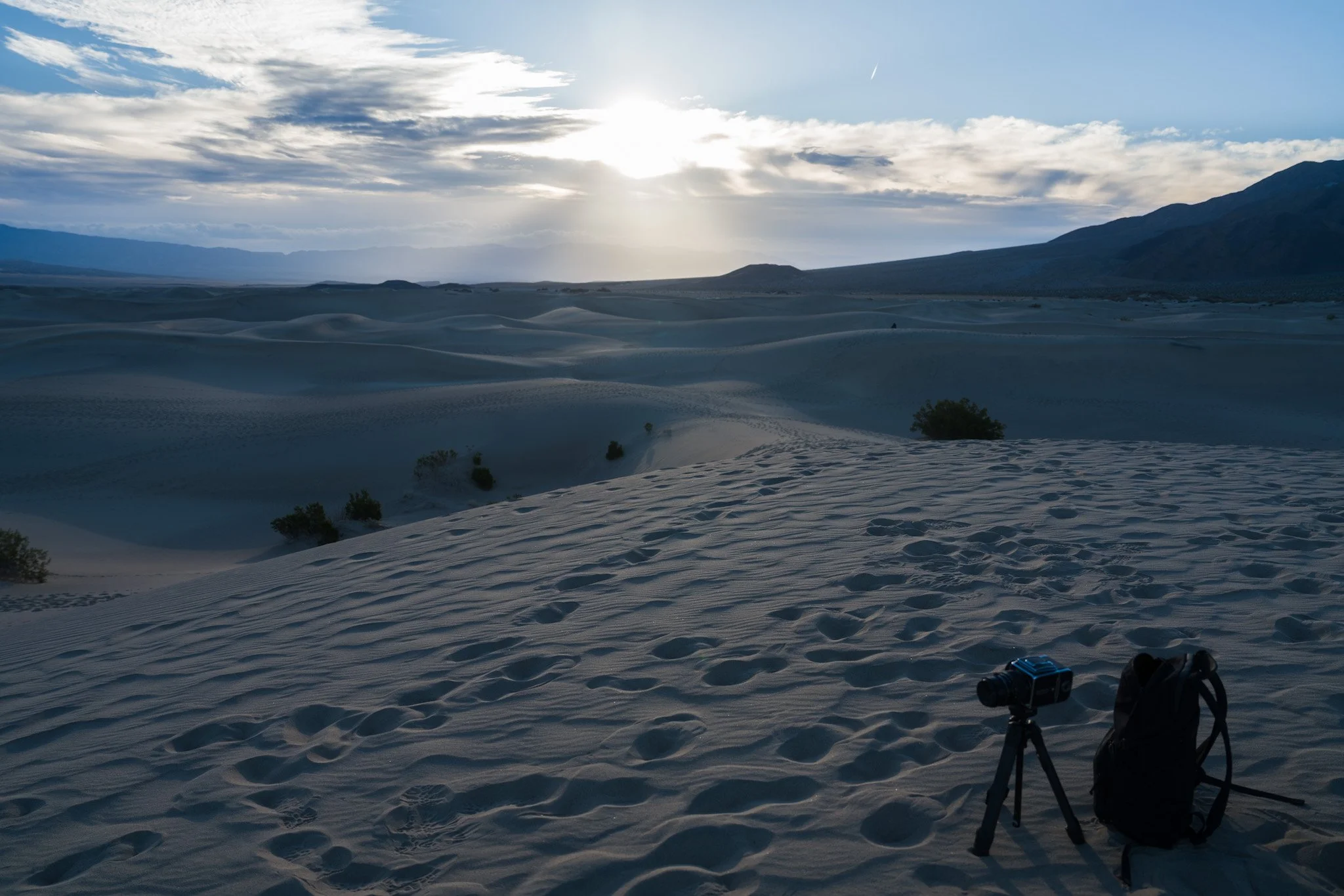 Desert landscape with sand dunes, some small bushes, distant mountains, cloudy sky, and a pair of binoculars on a tripod in the foreground.