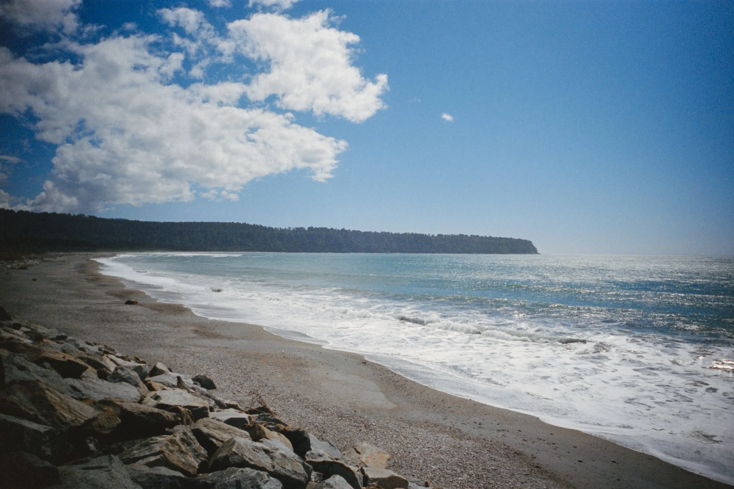 A scenic view of a beach with rocky shoreline, waves crashing, a forested cliff in the distance, and a bright blue sky with scattered white clouds.