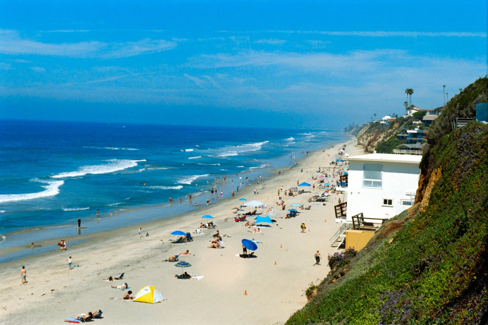 A crowded beach with umbrellas, people swimming, and houses on a hillside under a blue sky.