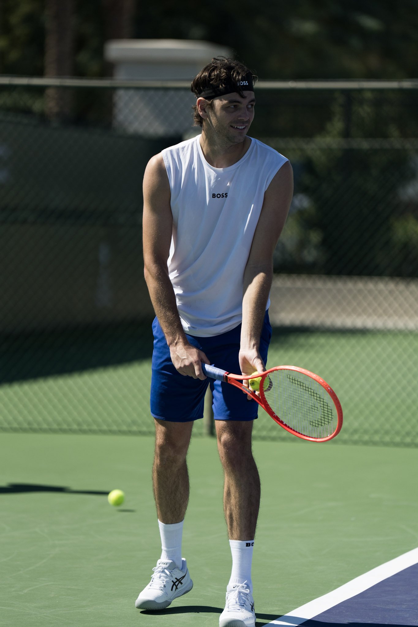A man playing tennis on a green outdoor court, wearing a white sleeveless shirt, blue shorts, white tennis shoes, white socks, and a black headband, with a tennis ball in front of him.