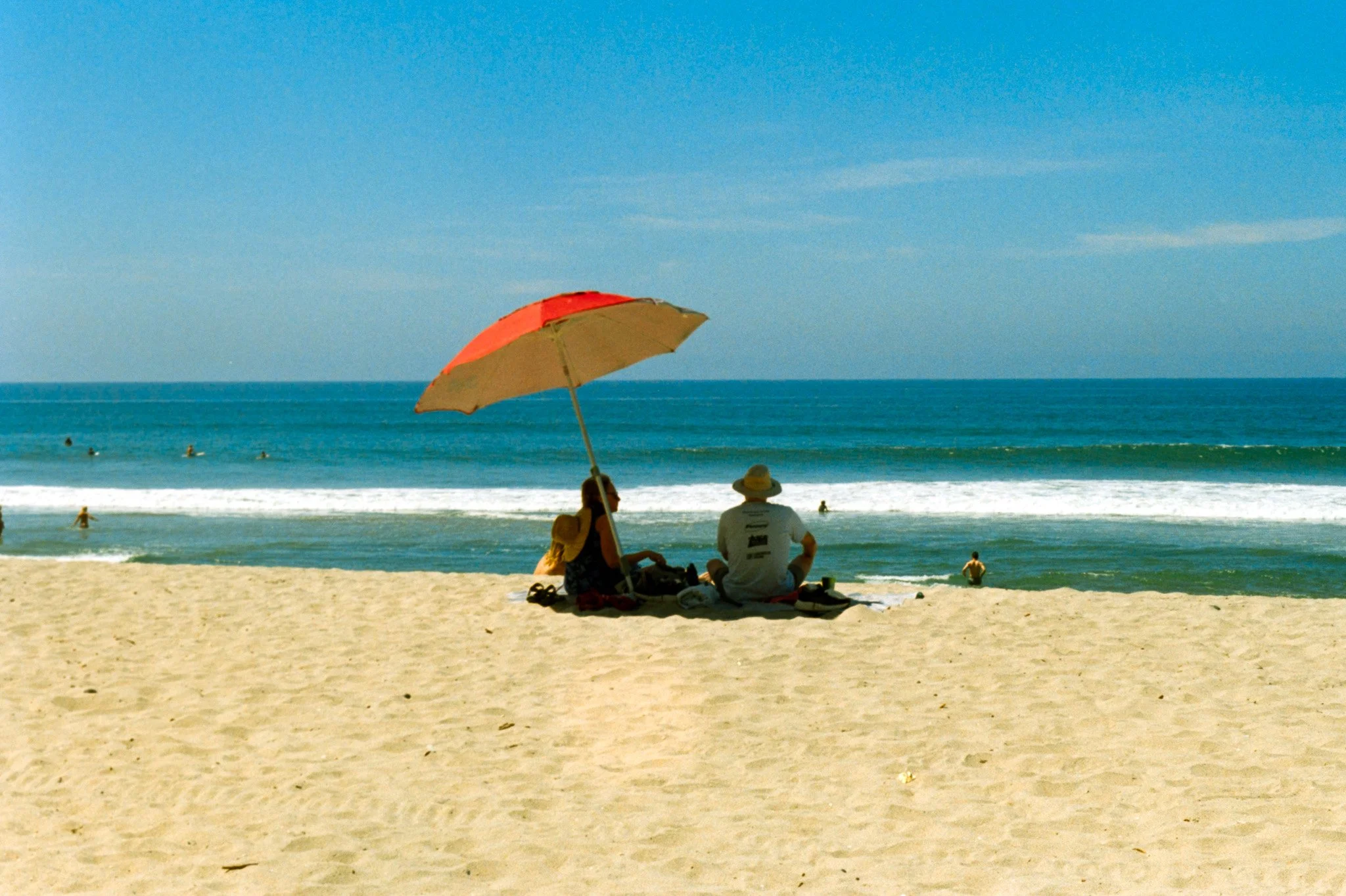 People sitting on a sandy beach under a red and yellow umbrella, facing the ocean with waves and a clear blue sky.