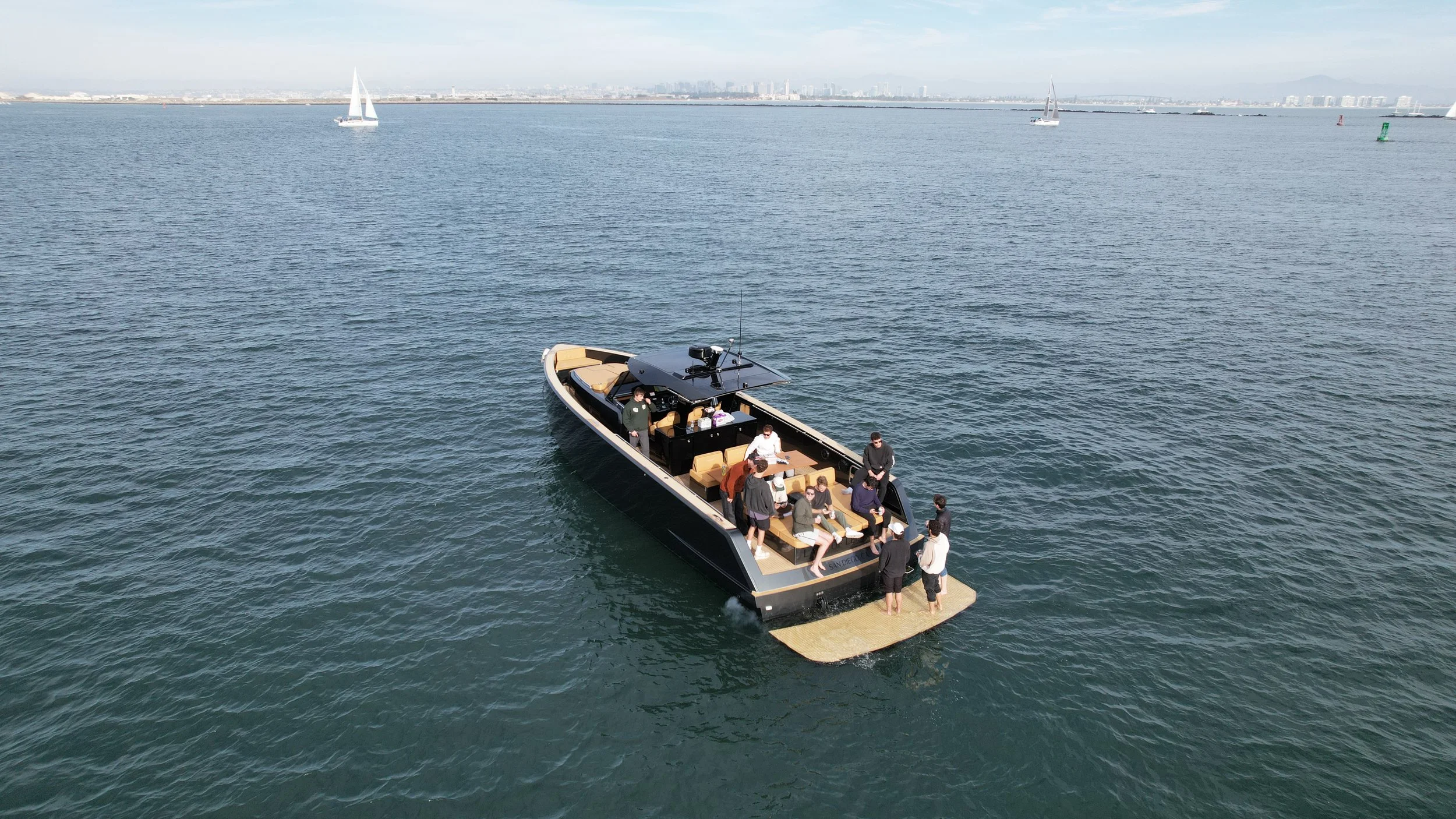 A group of people socializing on a yacht in the water with sailboats and a distant city skyline in the background.