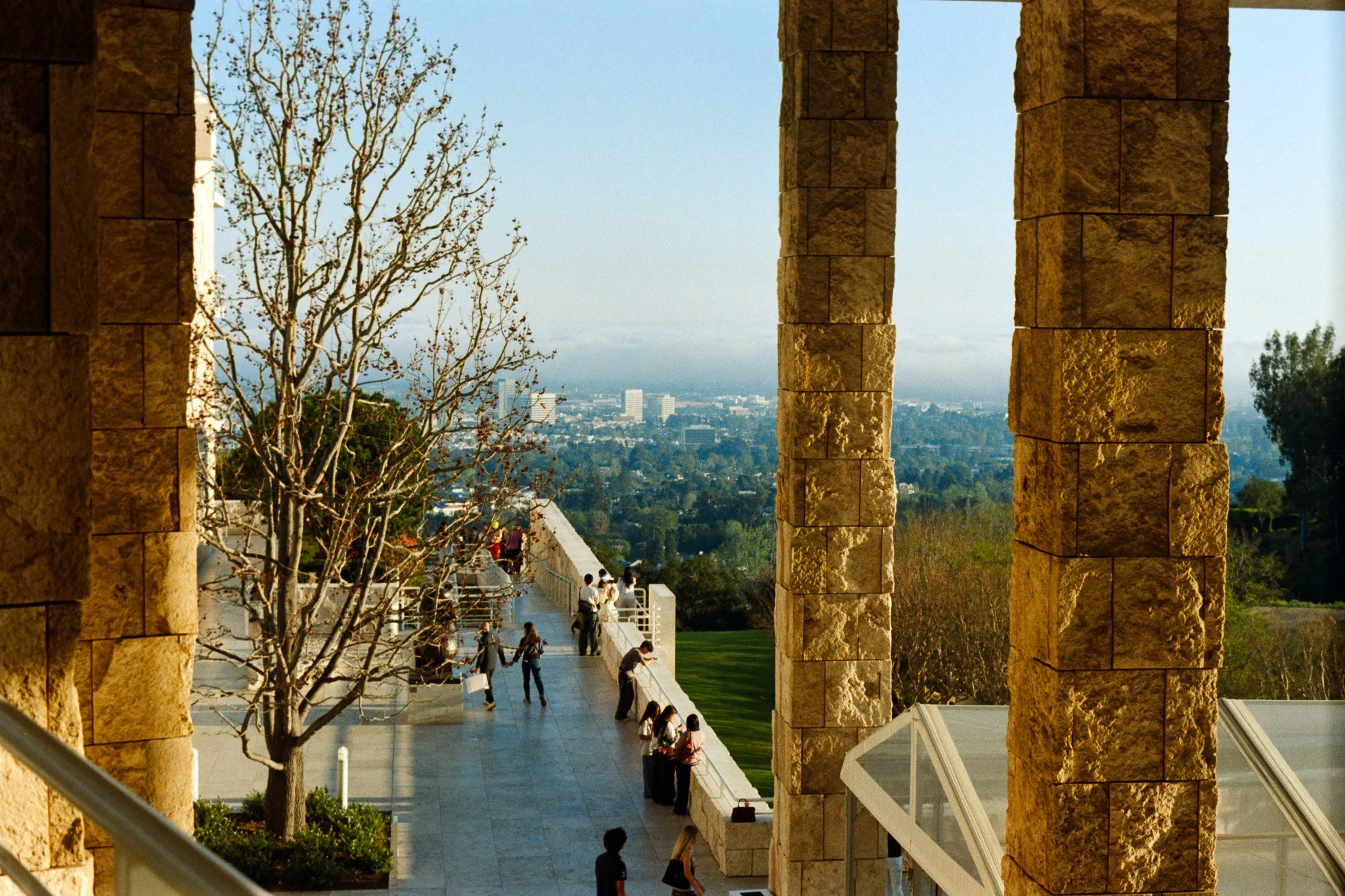 View through large stone archways showing a walkway with people, a tree, and a cityscape in the distance under a cloudy sky.