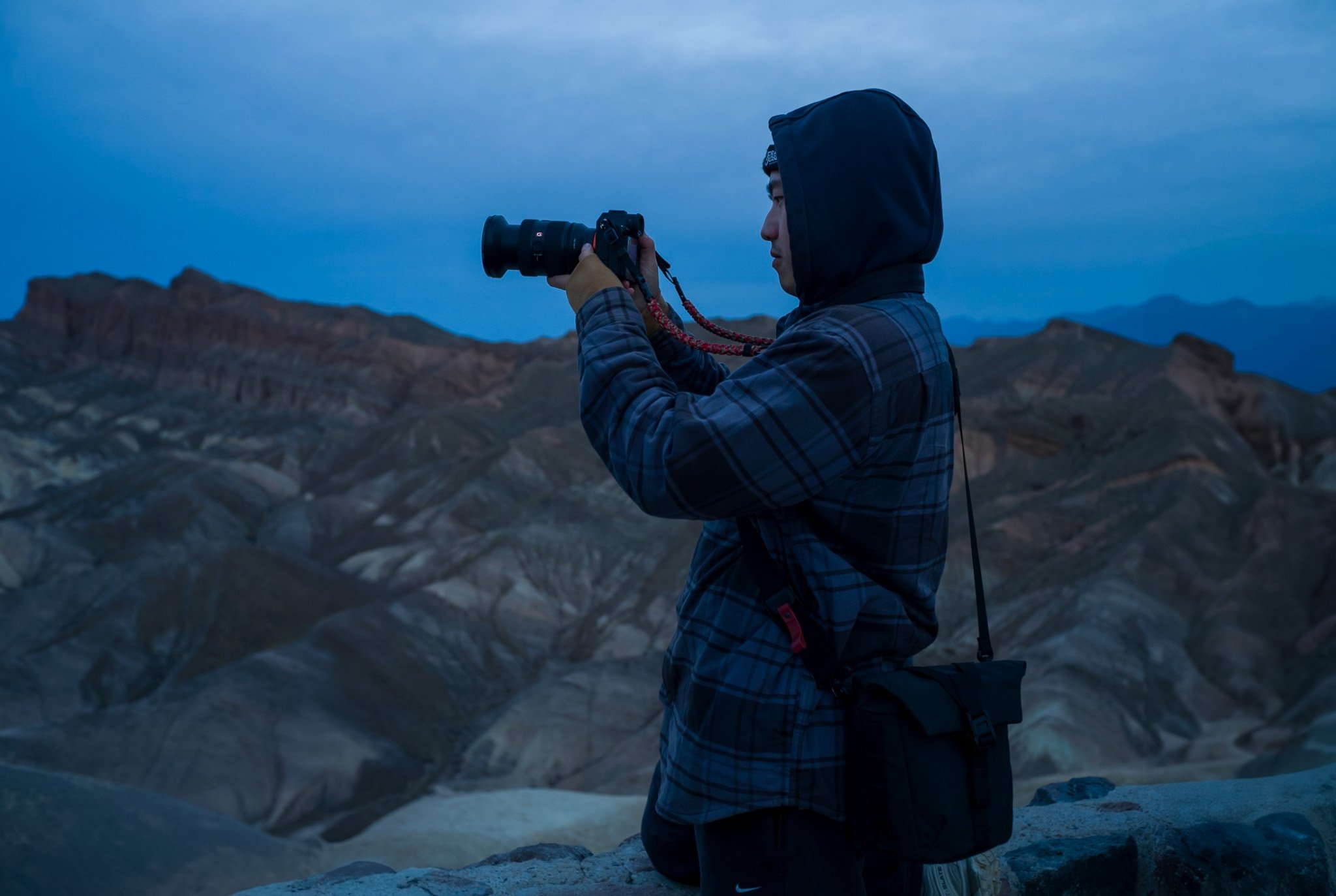 A person wearing a hoodie and plaid jacket taking a photograph with a camera in a mountainous, rugged landscape during dusk or dawn.