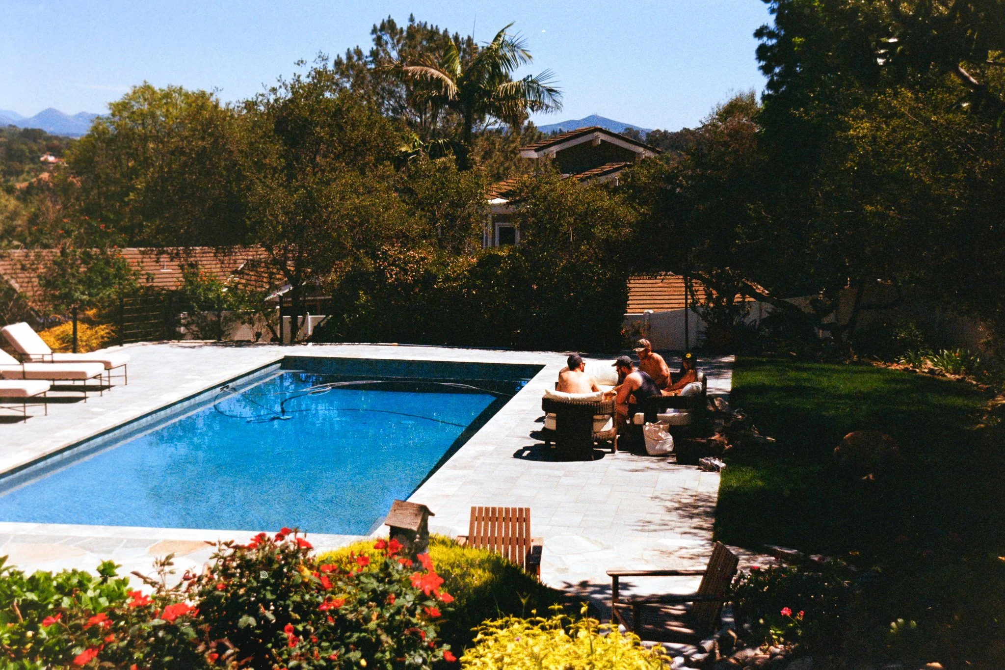 People sitting by a swimming pool on a sunny day, surrounded by trees and flowers, with a house and mountains in the background.