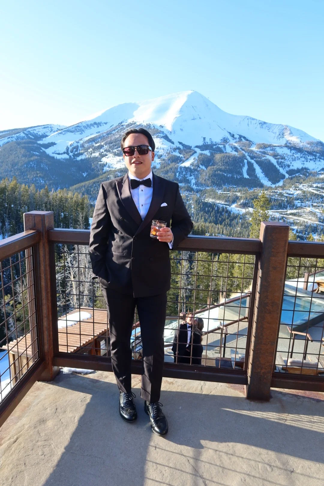 A man in a tuxedo and sunglasses standing on a balcony with a mountain and snowy landscape behind him, holding a glass of whiskey.