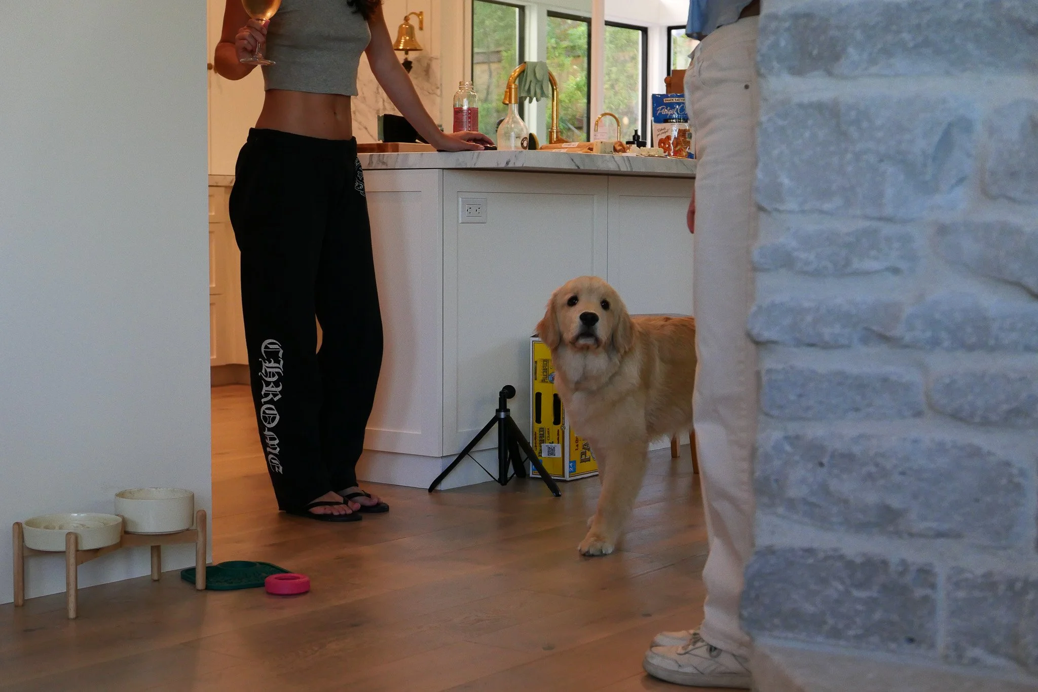 A woman with a gray tank top and black sweatpants holding a wine glass stands in a kitchen, with a golden retriever looking up at her. Part of a person in light-colored pajamas is visible on the right. The kitchen has a marble countertop, large windows, and a stone wall. There are dog bowls and a food container on the floor.