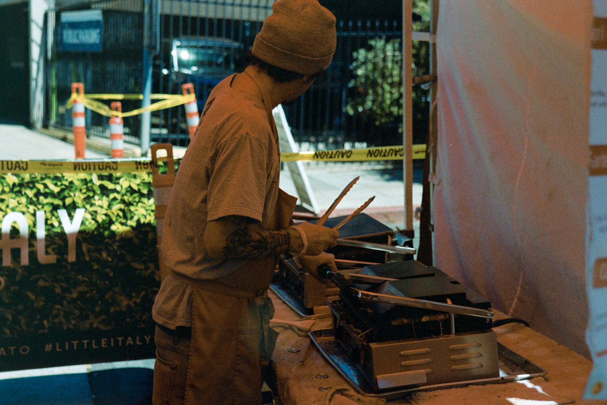 A person wearing a brown beanie and brown shirt looking at a menu or electronic device at an outdoor food stand with caution tape in the background.