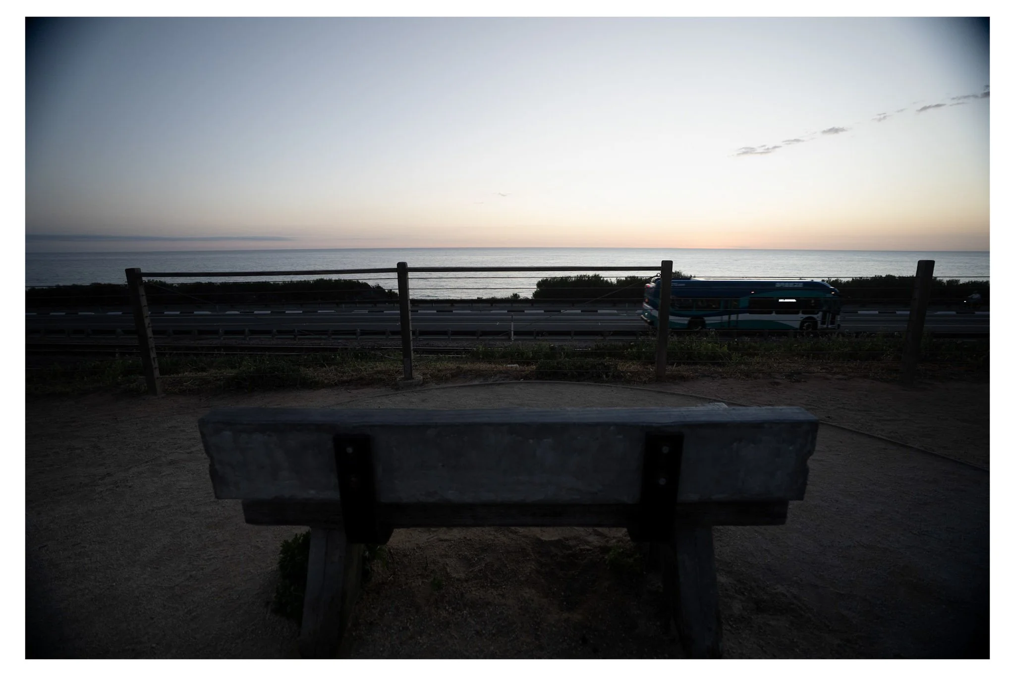 A view of the sunset over the ocean, with a bench in the foreground and a bus on the road.