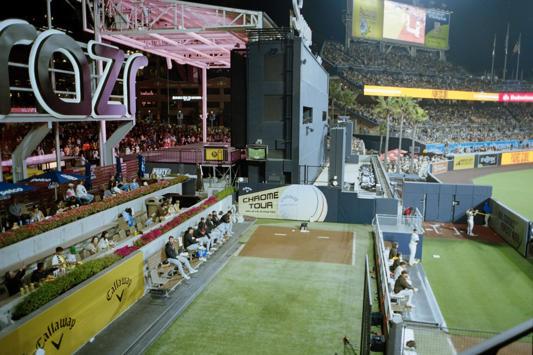 Nighttime view of a baseball stadium with players on the field and spectators in the stands. The image shows the dugout area with sitting players, advertisements on the fences, and a large scoreboard in the background.