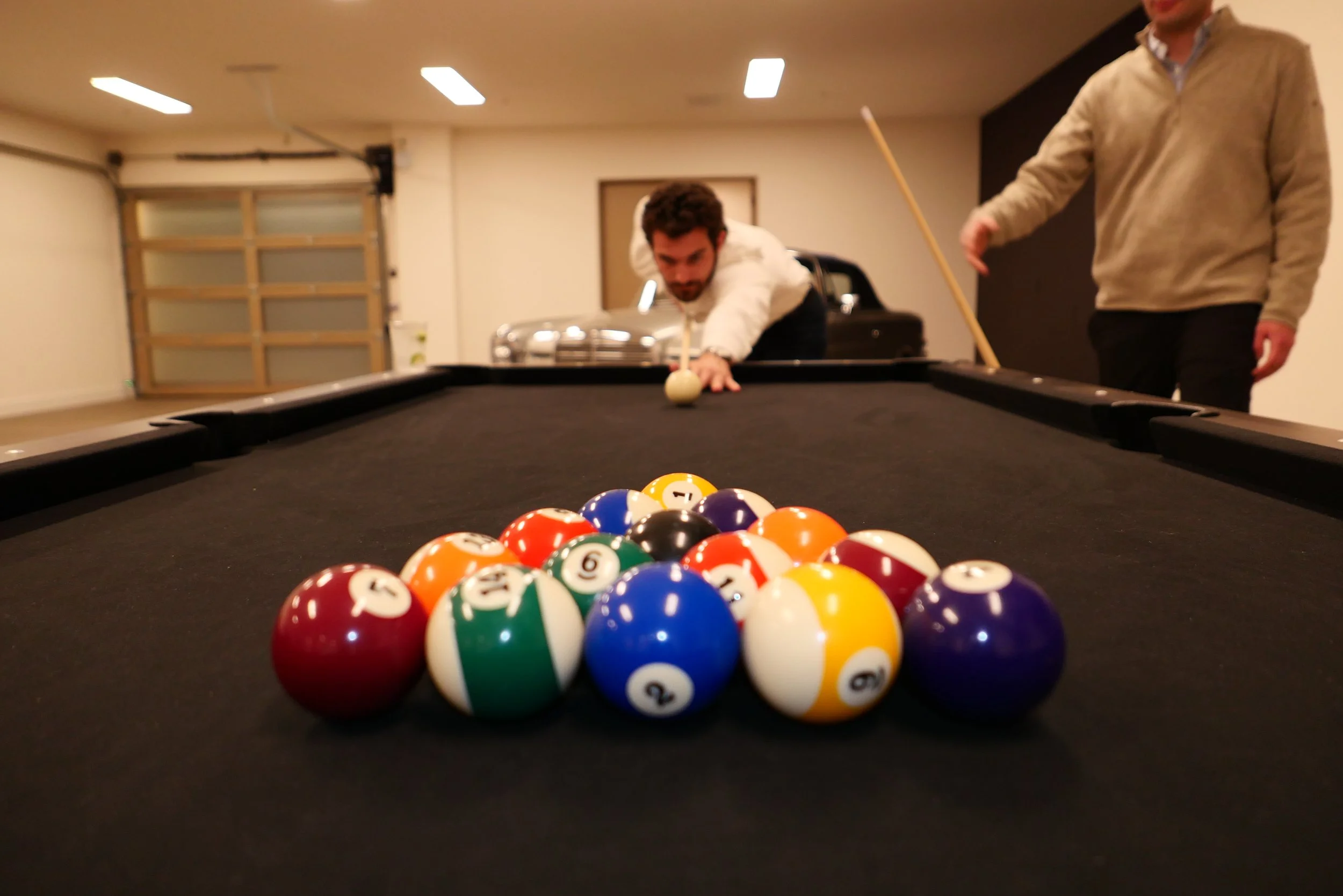 A pool table with billiard balls arranged in a triangle, with two men in the background preparing to play. One man is leaning over the table aiming to hit the cue ball, while the other stands nearby holding a cue stick.