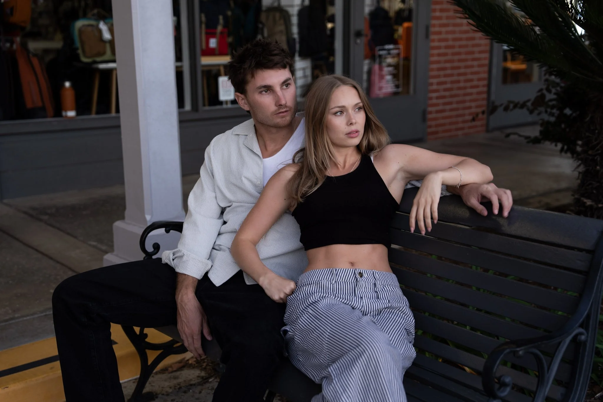A man with dark hair and a woman with light brown hair sitting on a park bench outside a store, with the woman leaning back and resting her arm on the bench while the man leans in slightly behind her, both looking into the distance.