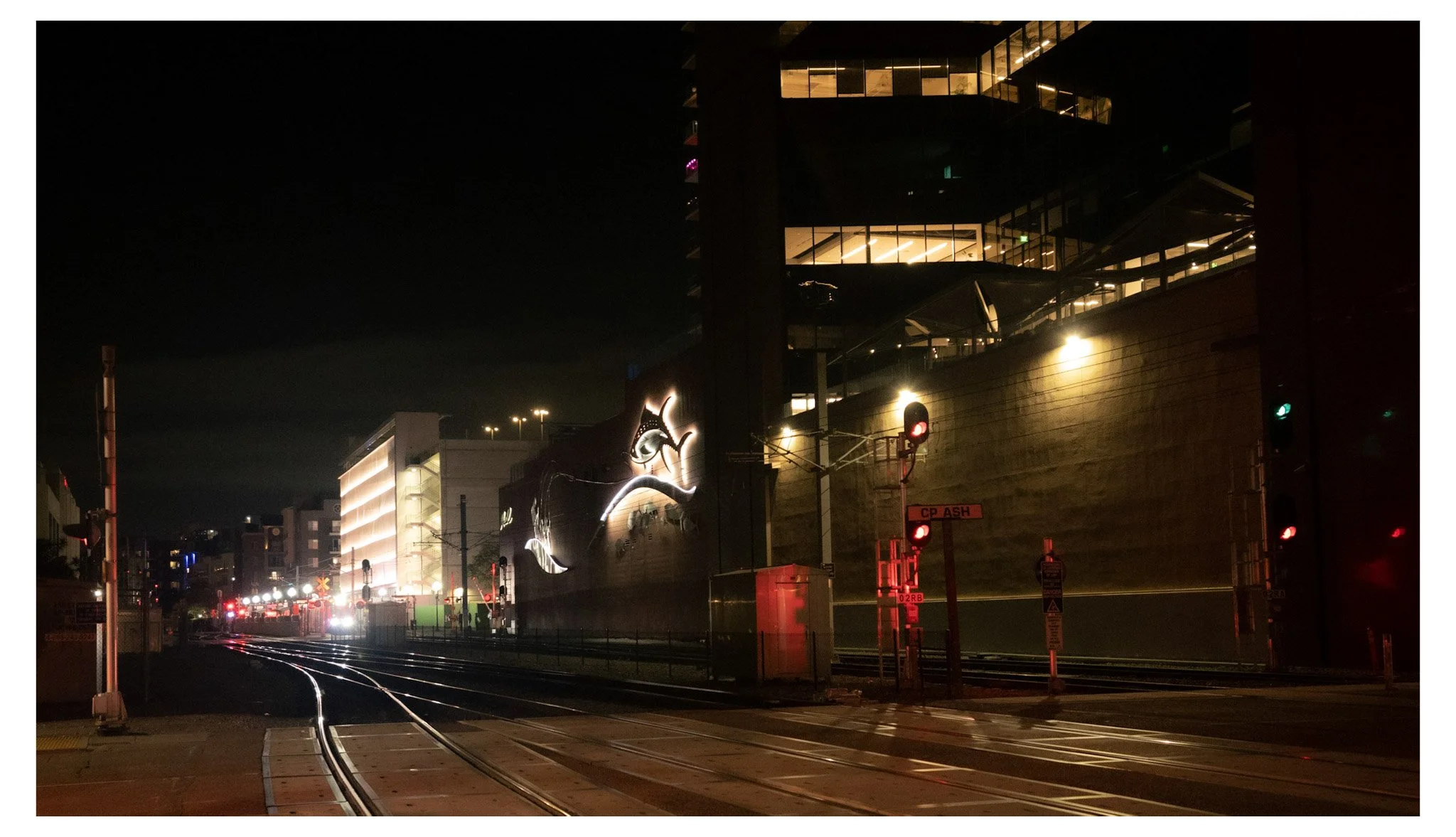 Nighttime scene of a modern city with illuminated buildings, traffic signals, and train tracks in the foreground.