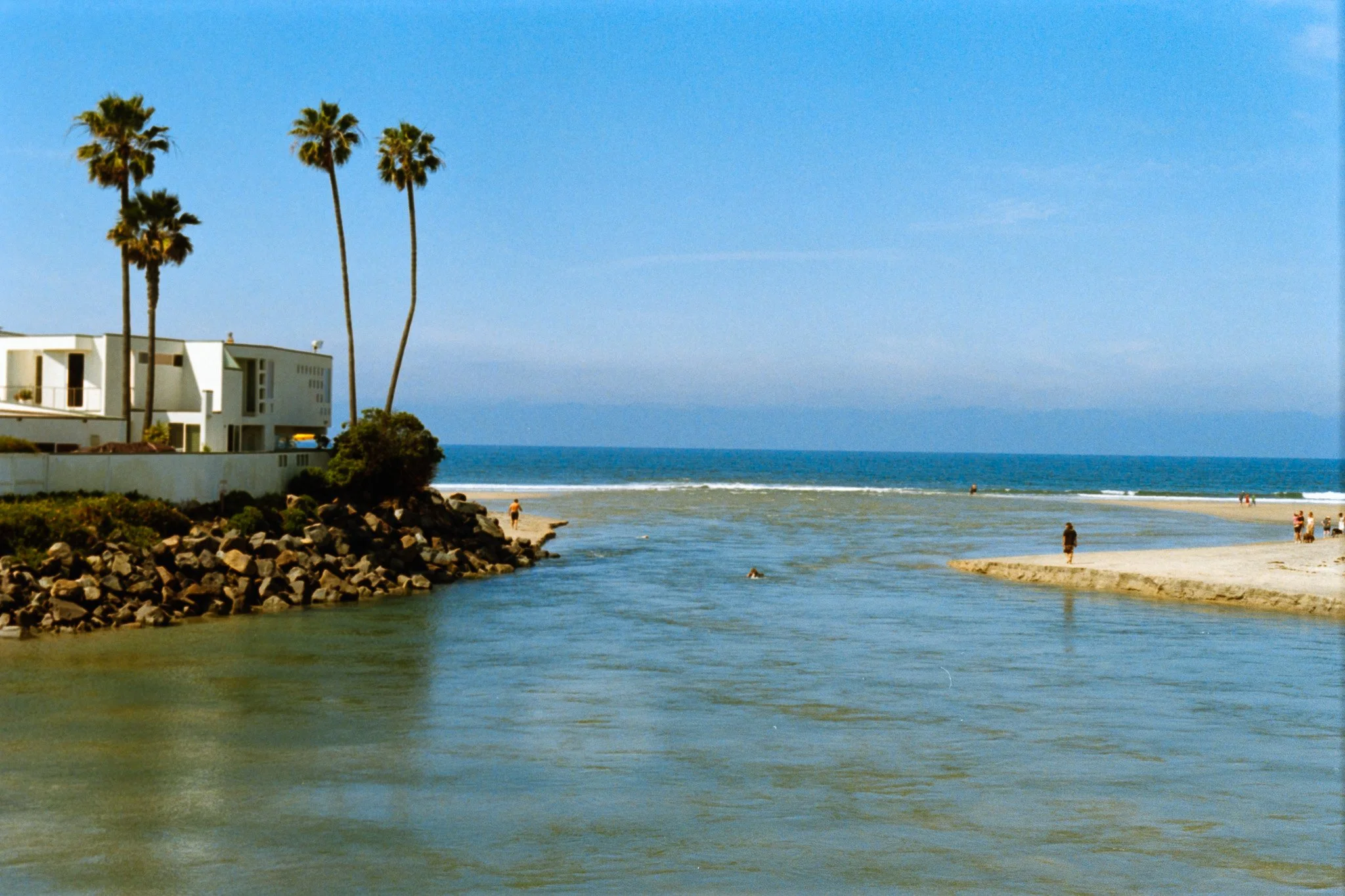 Beach scene with calm water, sandy shore, palm trees, a white modern building, and people walking or relaxing in the distance under blue sky.