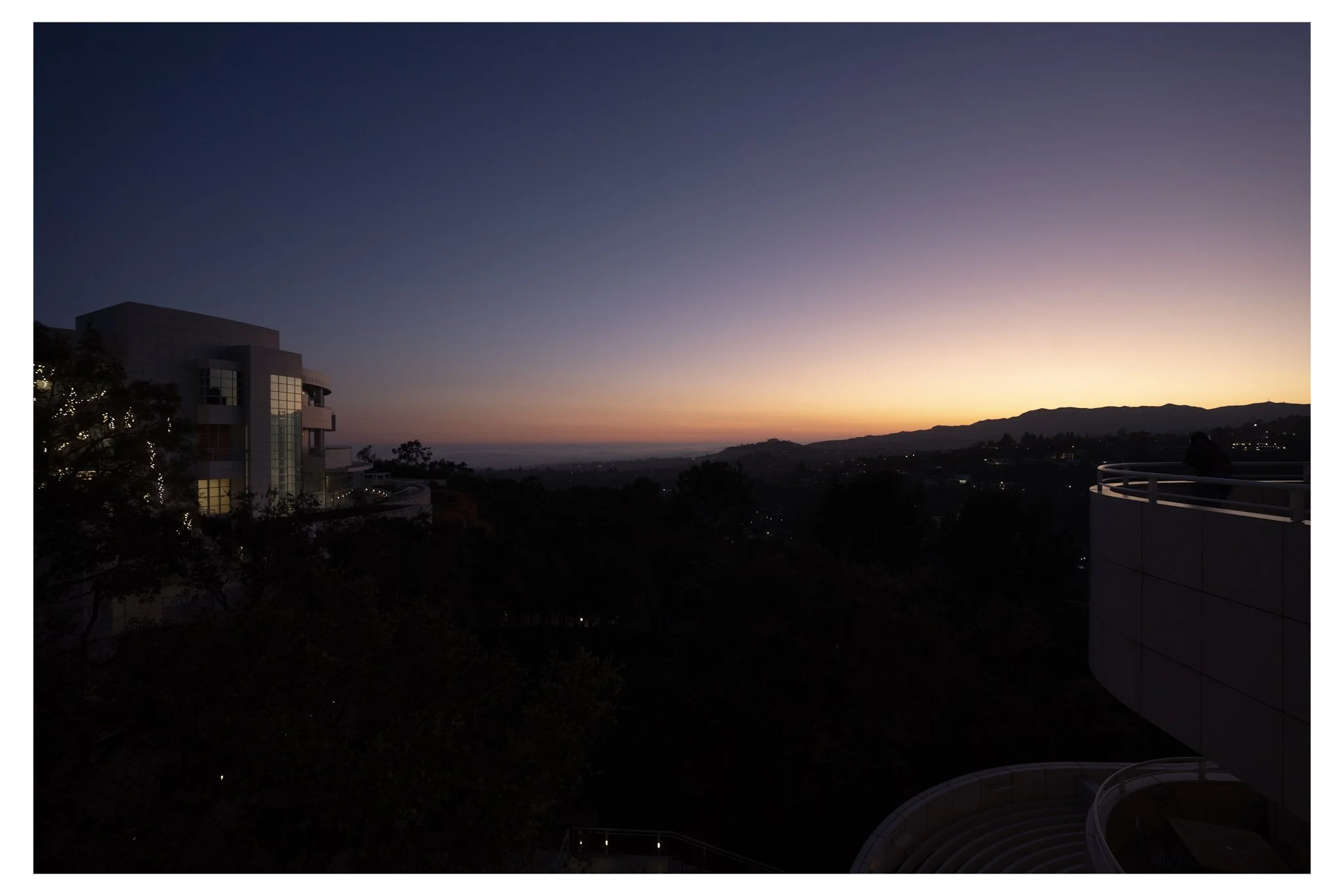 A sunset view with a building on the left, trees with evening lights, distant mountains, and a clear sky with fading sunlight.