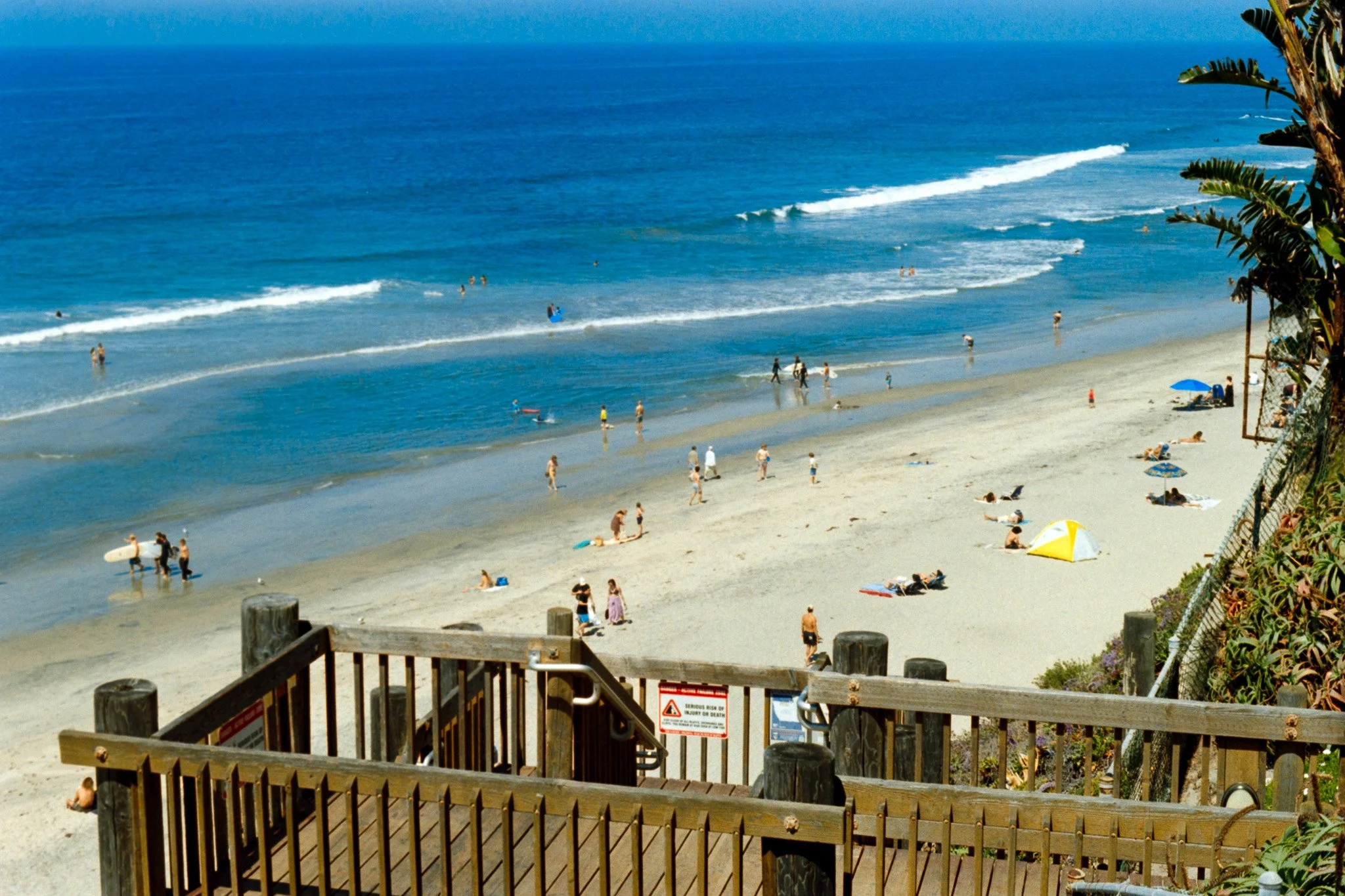 Beach scene with beachgoers swimming, sunbathing, and walking on sandy shores; ocean with gentle waves; blue sky; trees and plants along the fence near the stairs leading to the beach.