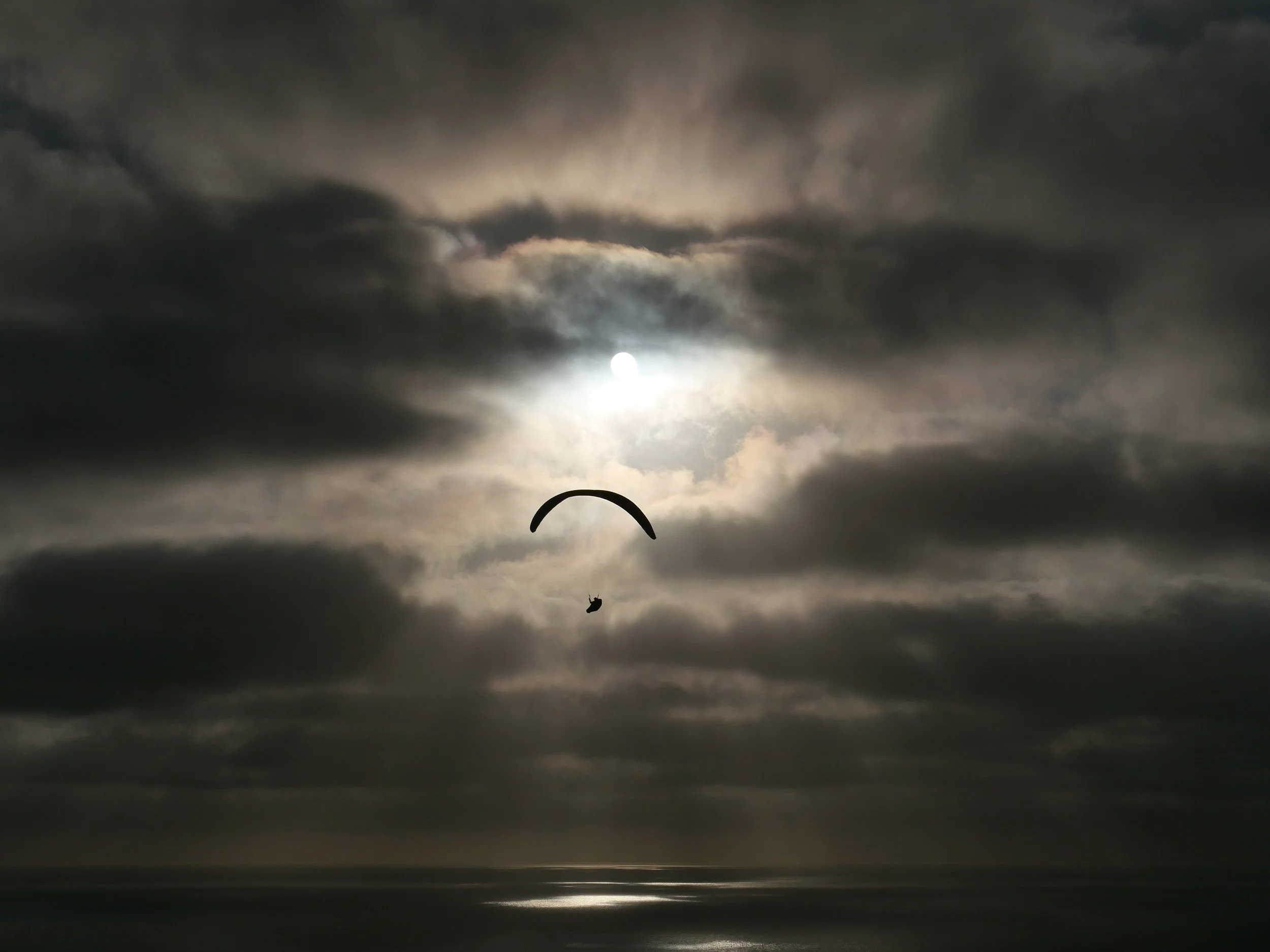 The image shows a night sky with dark clouds and the moon partially visible behind the clouds. A small silhouette of a paraglider is seen in the foreground, flying beneath the cloudy sky.