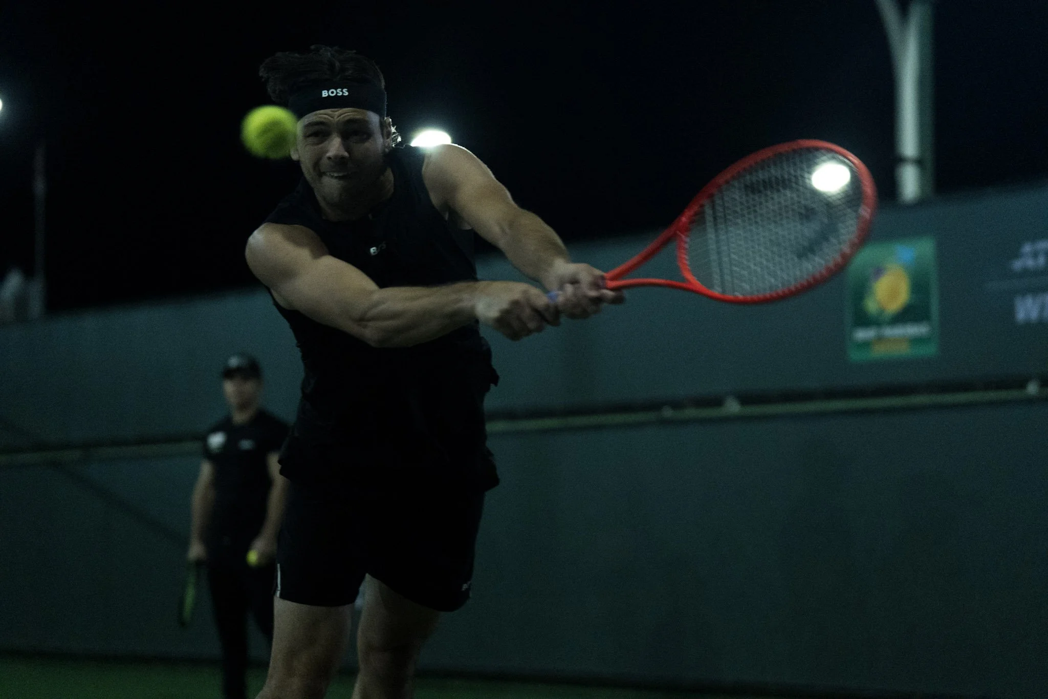 A male tennis player hits a yellow tennis ball with a red racket on an indoor court at night, with a dark background and another player in the distance.