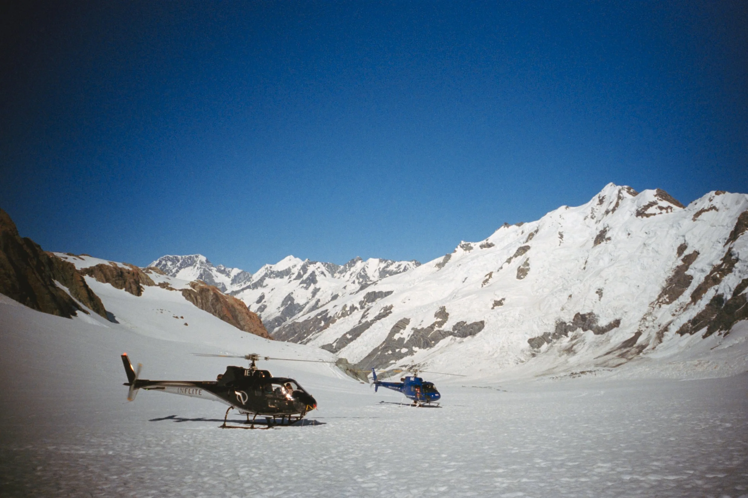 Helicopters on snow-covered landscape with snow-capped mountains in the background under a clear blue sky.
