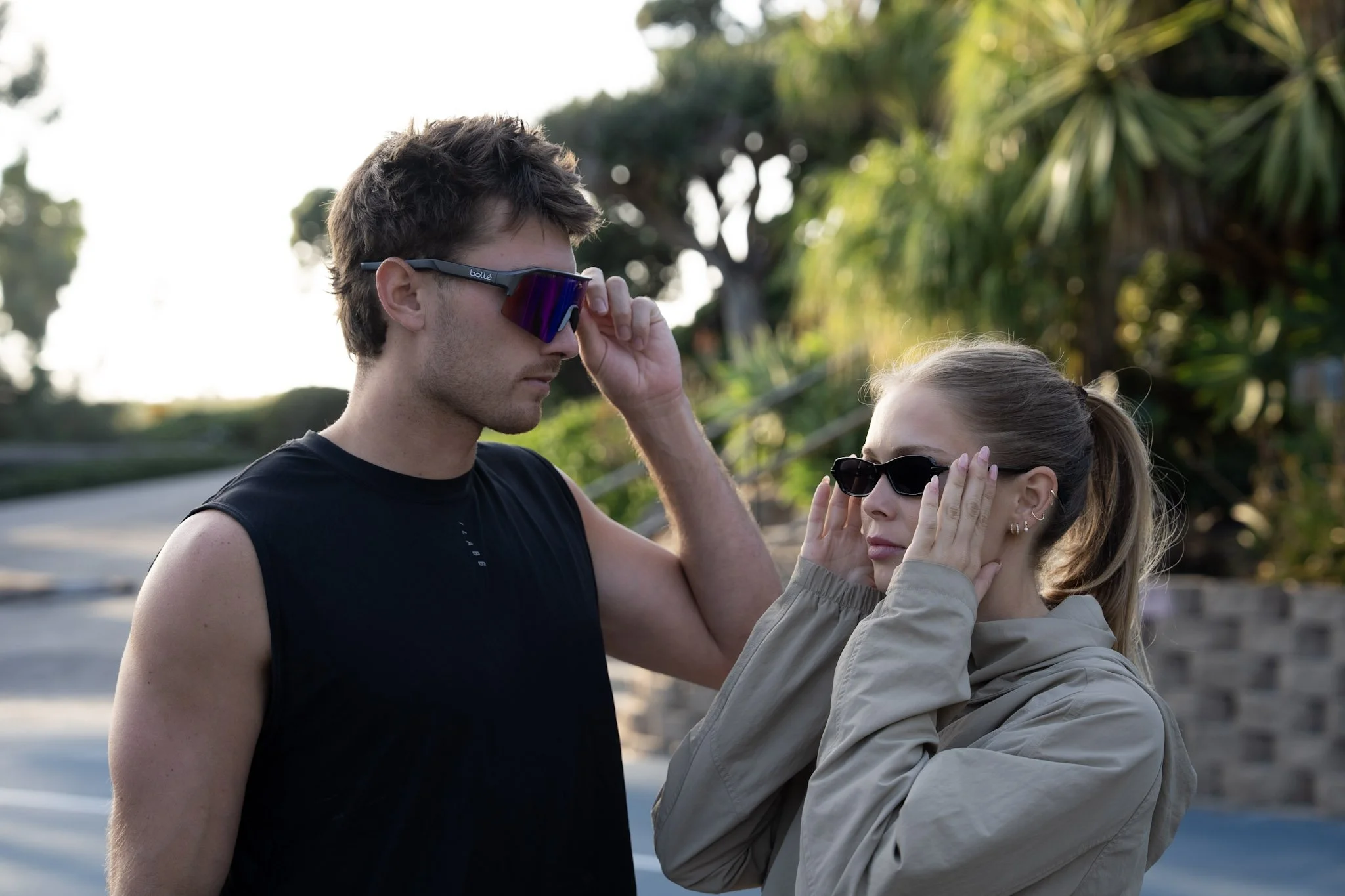 A man with short, dark, slightly messy hair and sunglasses is adjusting his glasses as a woman with long blonde hair in a ponytail and sunglasses touches her face, both standing outdoors with blurred trees in the background.