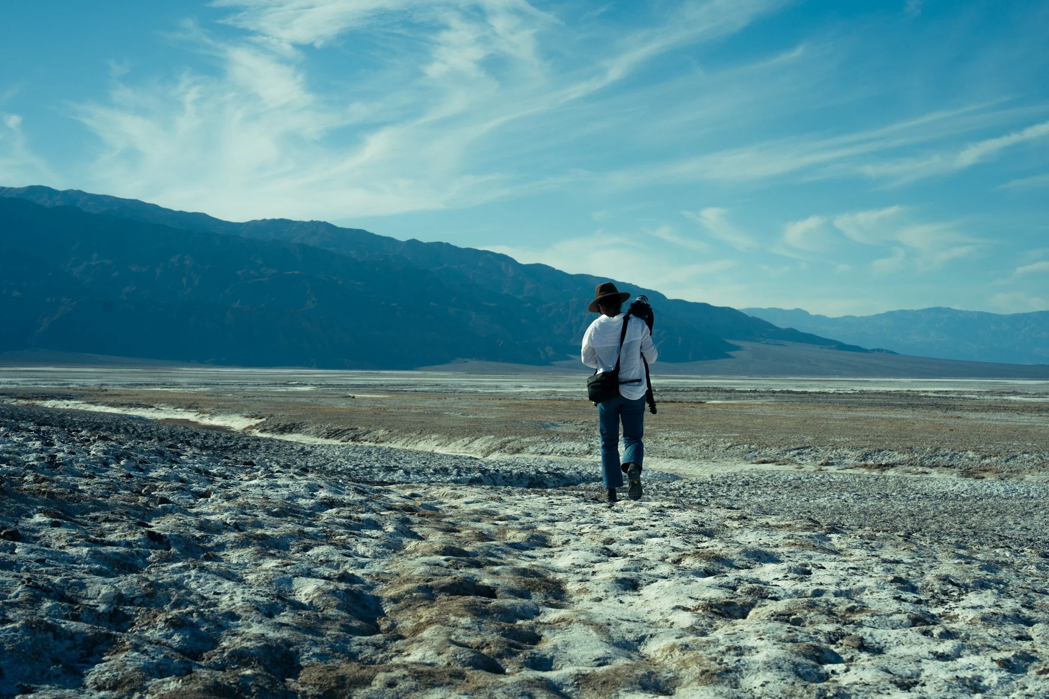 A person with a backpack, wearing a hat and jeans, walking in a barren landscape with mountains in the background and a partly cloudy sky.