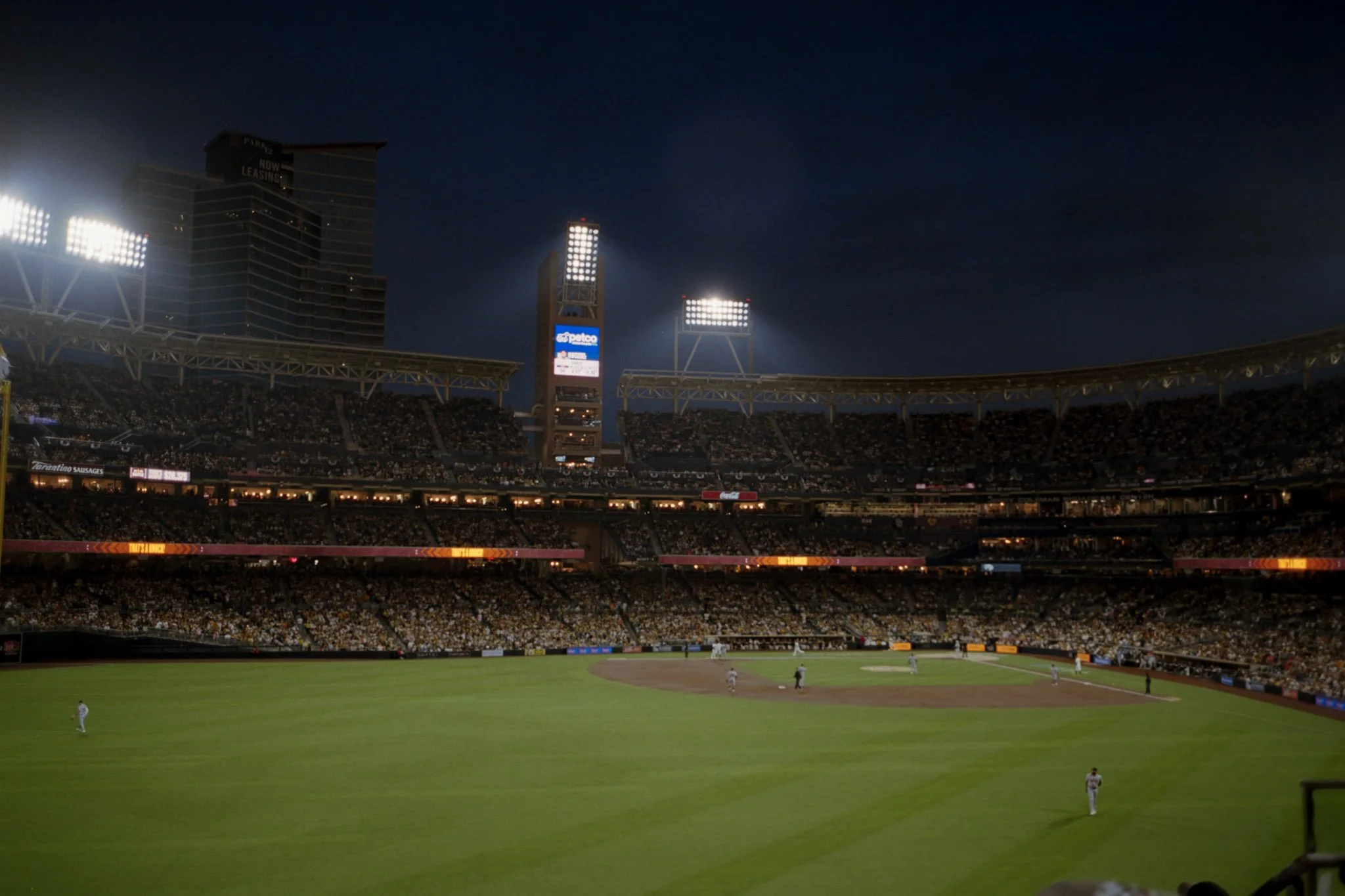A baseball game is being played at night in a large stadium with bright lights illuminating the field. The stands are filled with spectators, and players are on the field for the game.