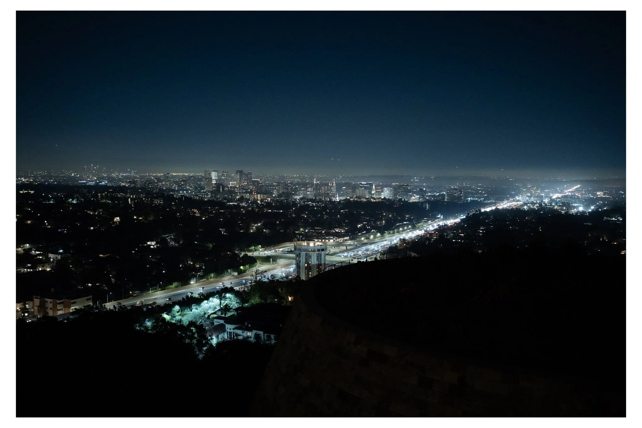 Nighttime cityscape view of a brightly lit urban area with high-rise buildings and highways from a hill or mountain.