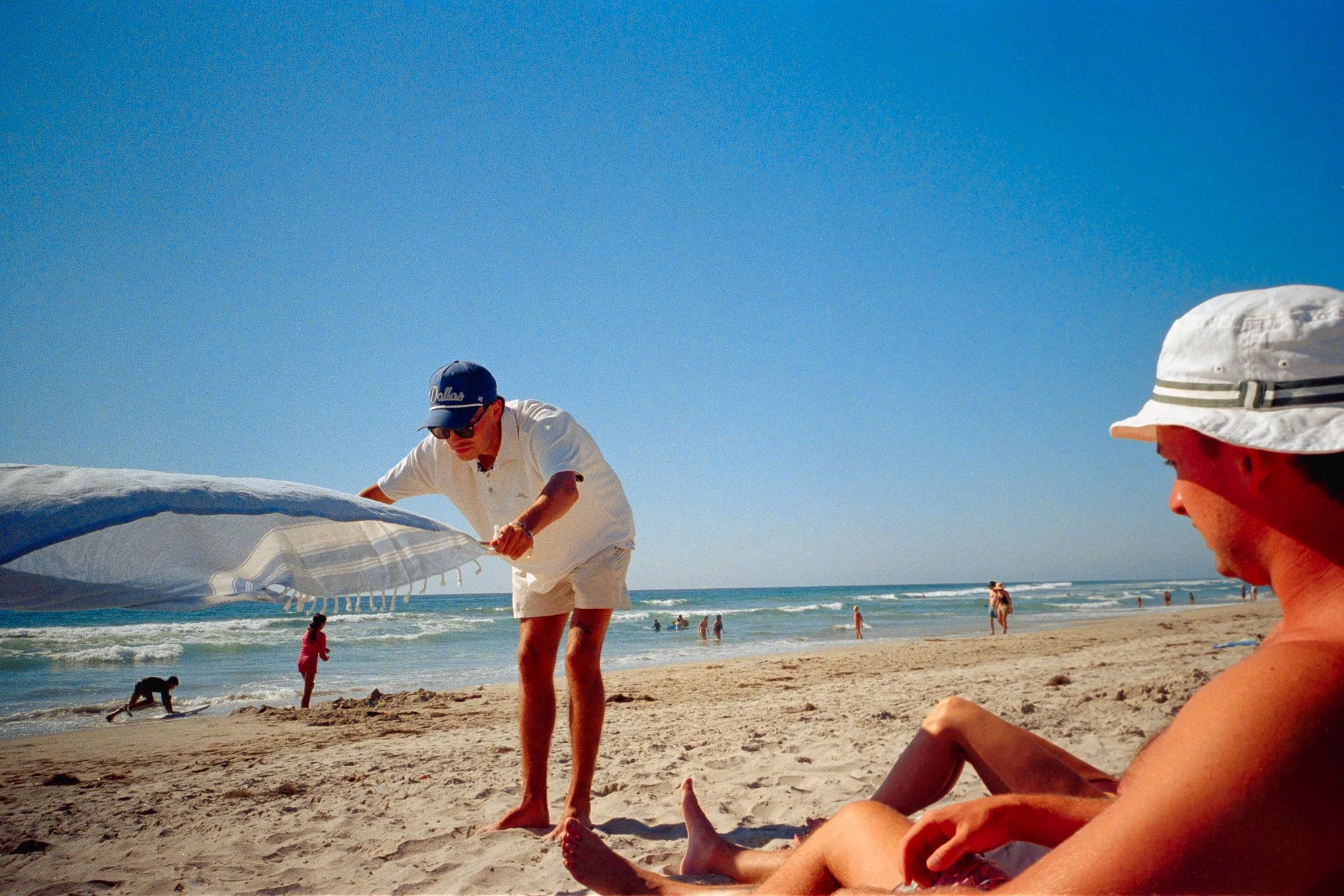 People at the beach, some children playing in the sand, and a man holding a windsurfing sail, with the ocean and clear blue sky in the background.