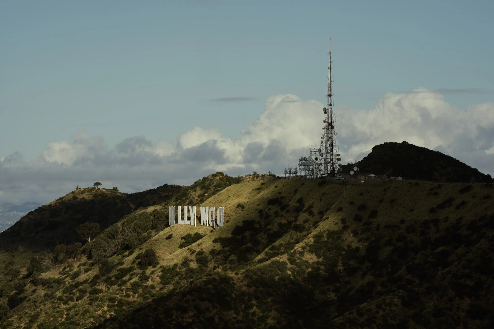 A hillside view featuring a large Hollywood sign and a communication tower with antennas on top, under a partly cloudy sky.