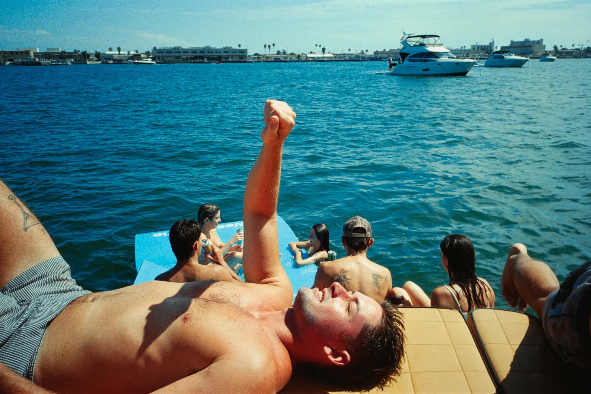 A group of people relaxing by the water on a boat, with boats anchored in the background and a shoreline with buildings and palm trees.
