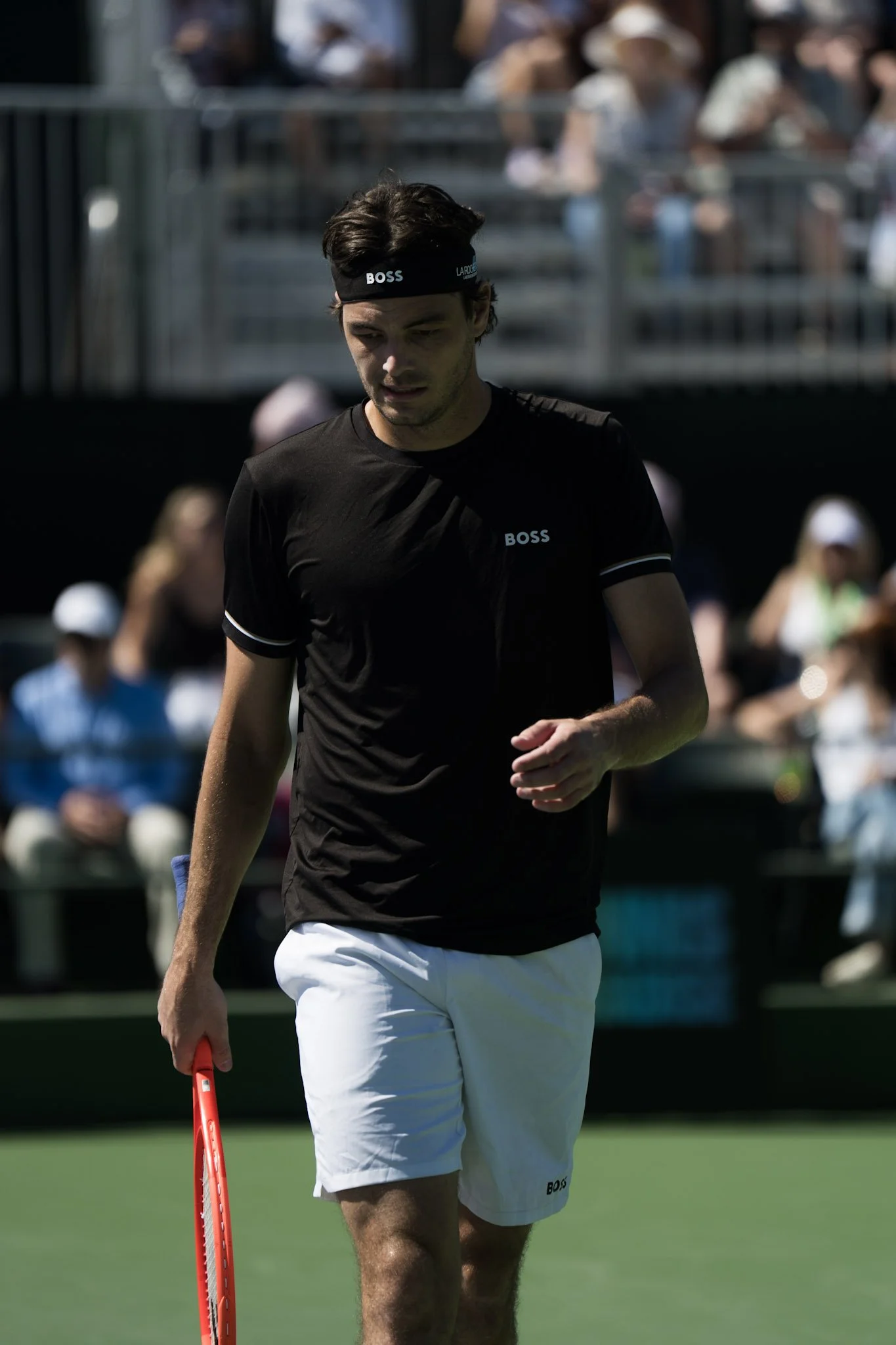 A male tennis player wearing a black shirt and white shorts, holding a tennis racket, walking on a tennis court during a match or practice session.