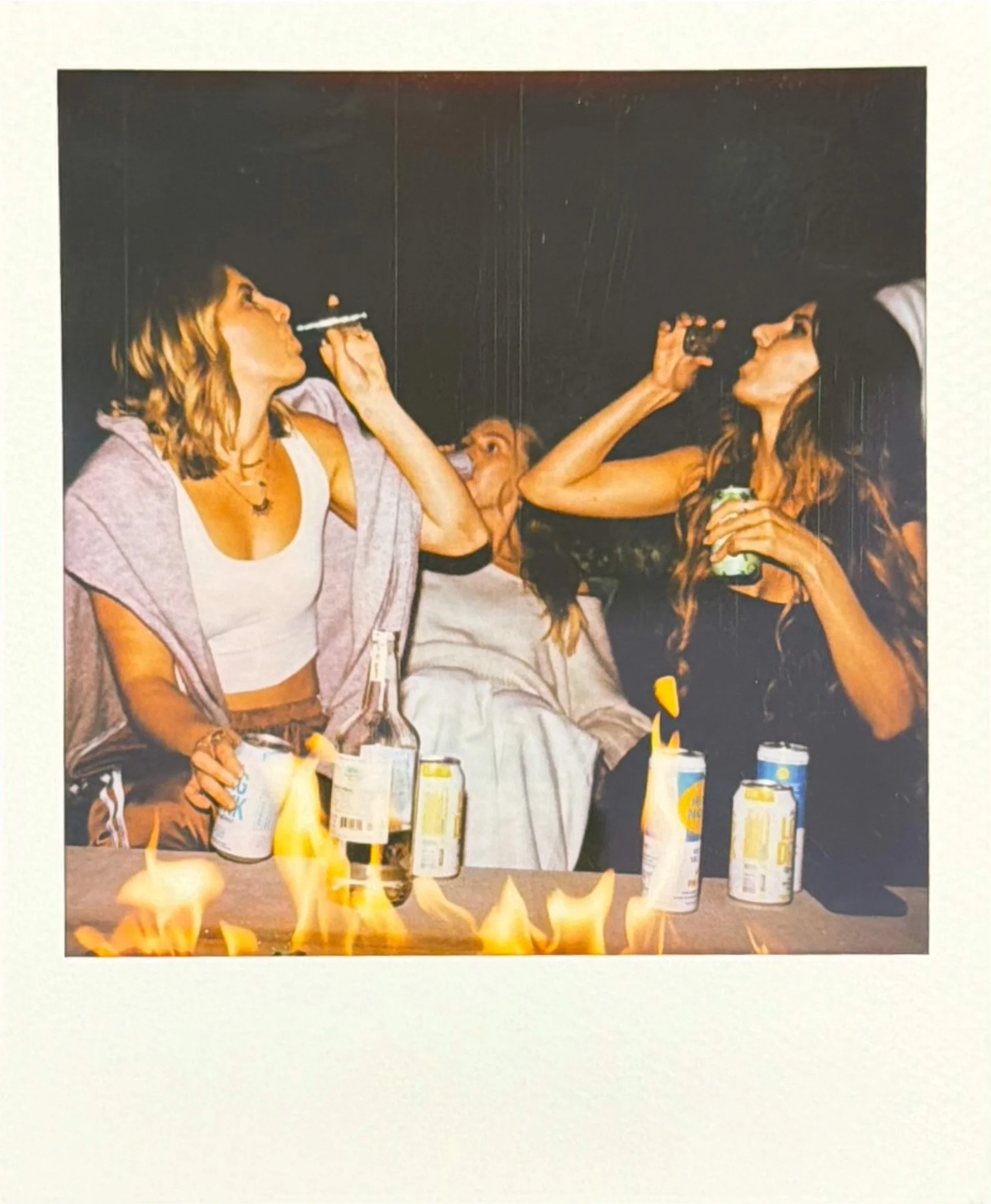 Two women with dark hair, sitting at a table, facing each other, holding drinks, and about to kiss. There are cans and bottles on the table, and a small fire on the table in the foreground.