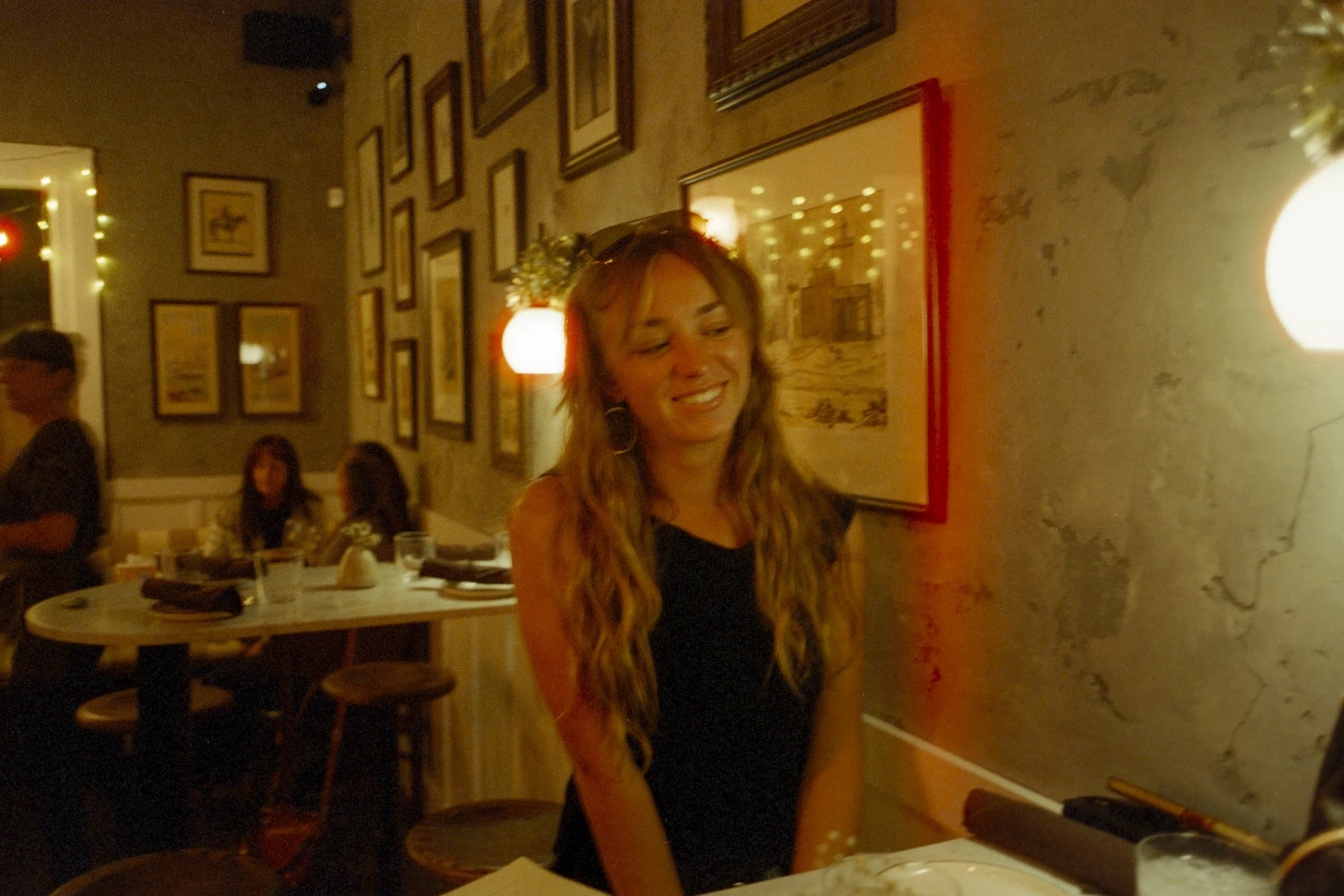 A woman with long wavy hair smiling at a restaurant table. The background shows wall decor with framed pictures and other patrons dining. Warm, dim lighting.