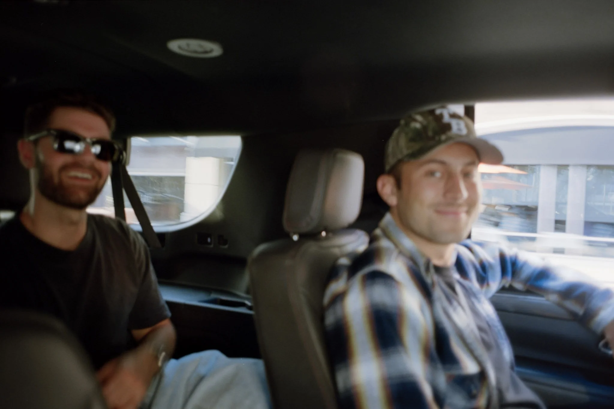 Two men sitting in the back seat of a car, smiling, with the man on the right wearing a plaid shirt and the man on the left wearing sunglasses.