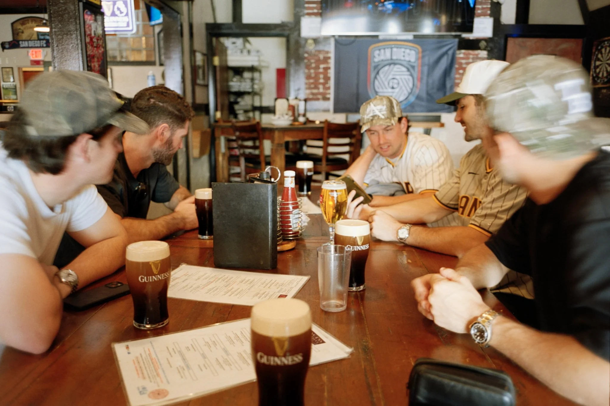A group of men sitting at a wooden table in a bar or restaurant, some wearing baseball caps, with glasses of Guinness beer, menus, and condiments on the table. The background shows a brick wall, a dartboard, and a banner with the logo 'San Diego'.