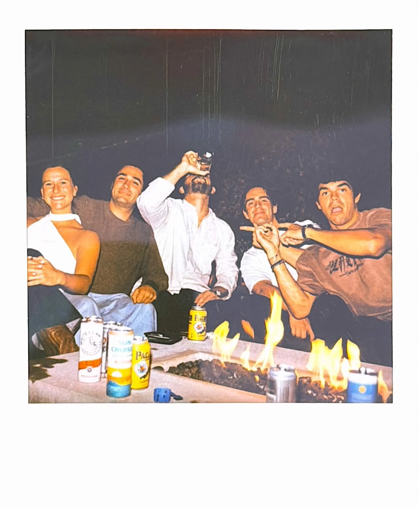 Group of five people sitting around a barbecue grill with flames. The table has various cans, including beers and a yellow can. The people are smiling, and one person is raising a glass. The background is dark.