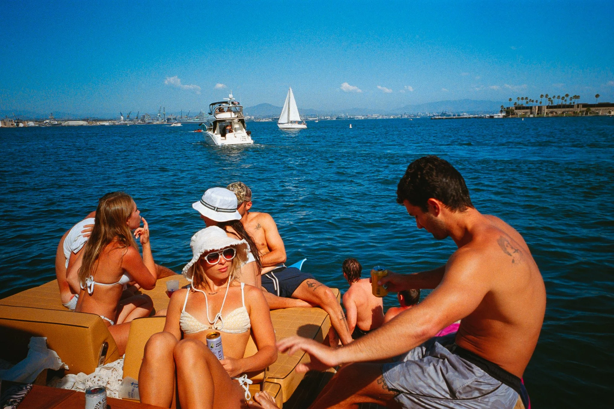 Group of people relaxing on a boat with a body of water, sailboats, and a cityscape with palm trees in the background.