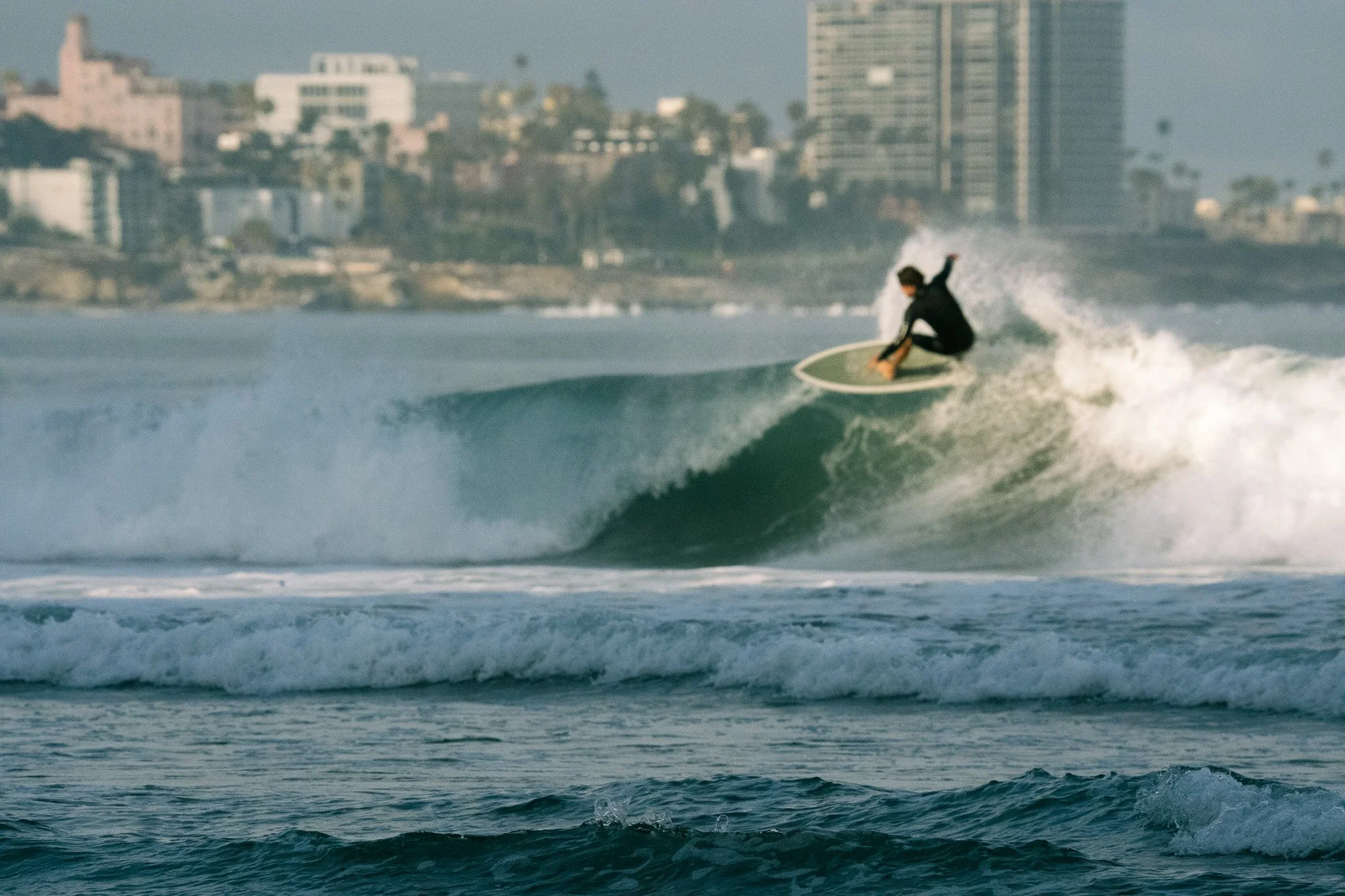 A person surfing on a wave in the ocean near an urban coastline with high-rise buildings in the background.