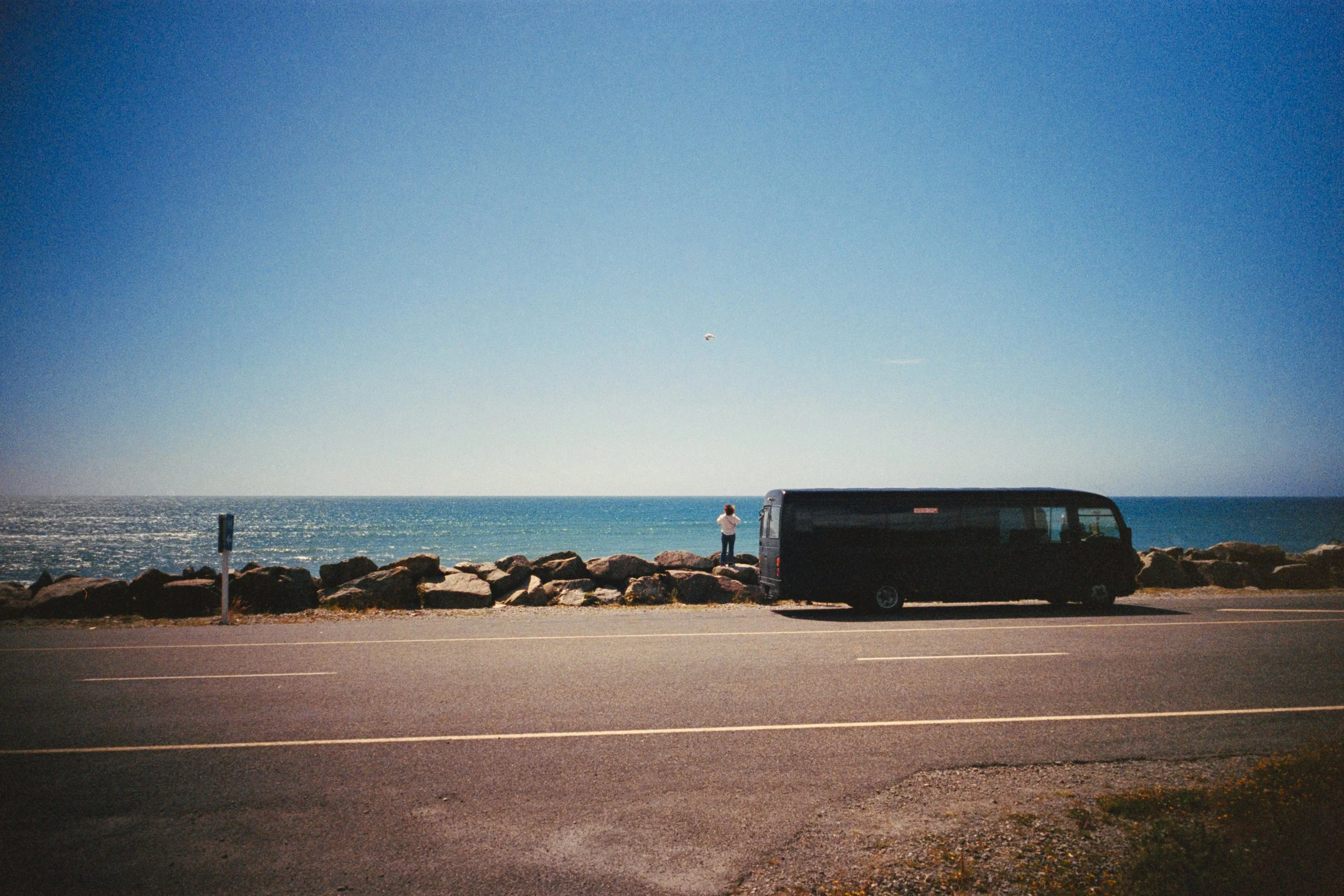A person standing by a black van on the side of a road overlooking the ocean, with rocks along the shoreline and a clear blue sky, at what appears to be a coastal location.