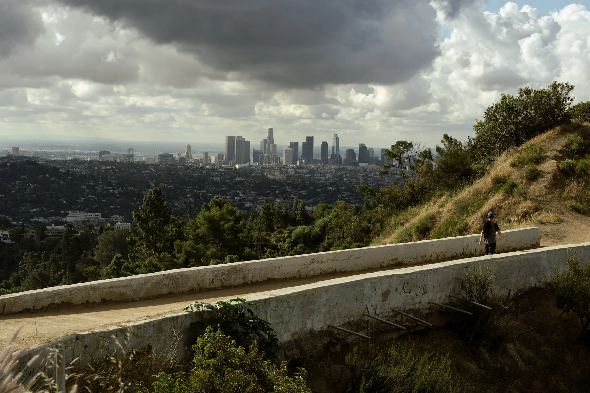 A hilly trail with a person walking, overlooking the city skyline of Los Angeles, under a cloudy sky.