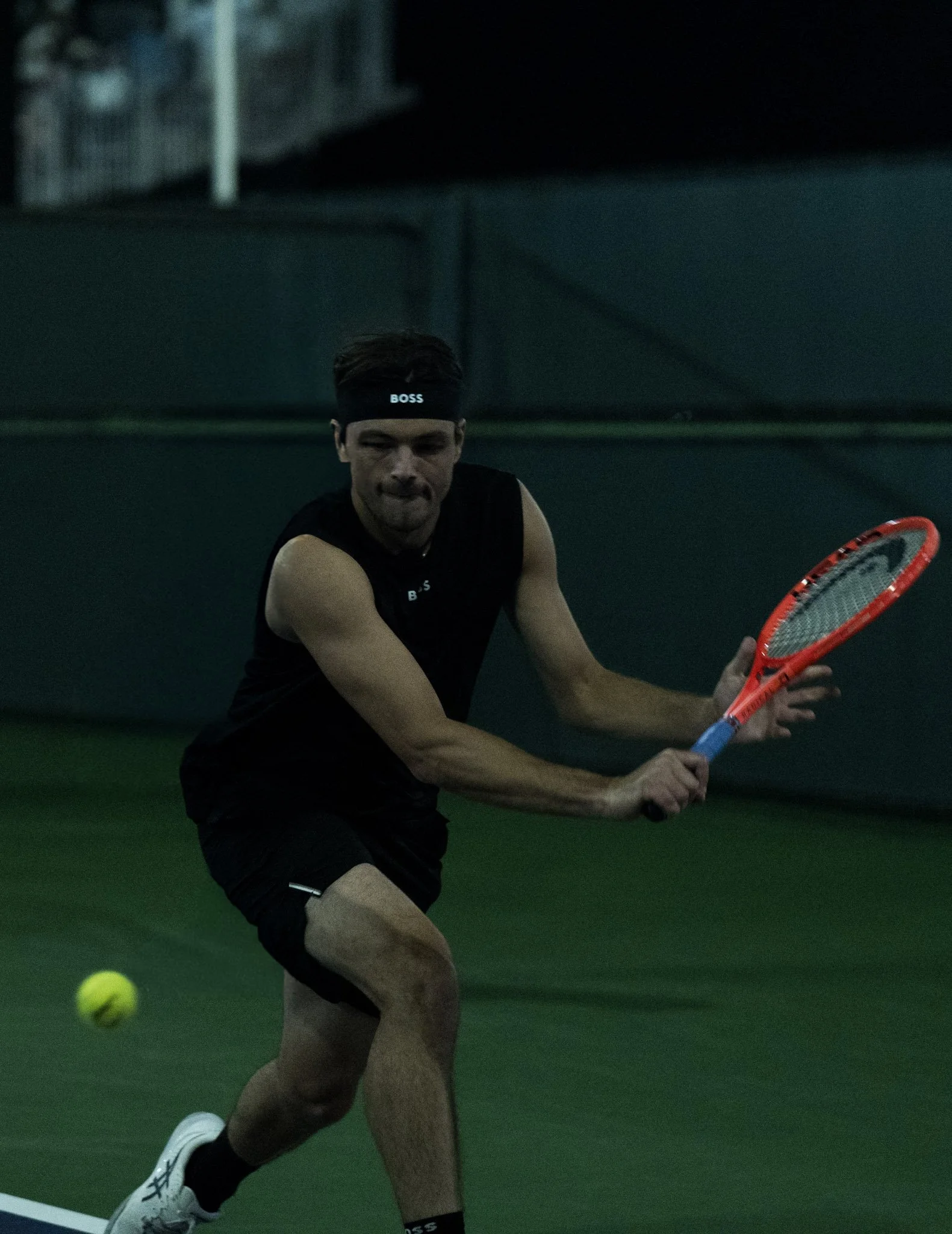 Tennis player in black athletic clothing and a headband labeled "BOSS", preparing to hit a tennis ball with an orange tennis racket on a green indoor court.
