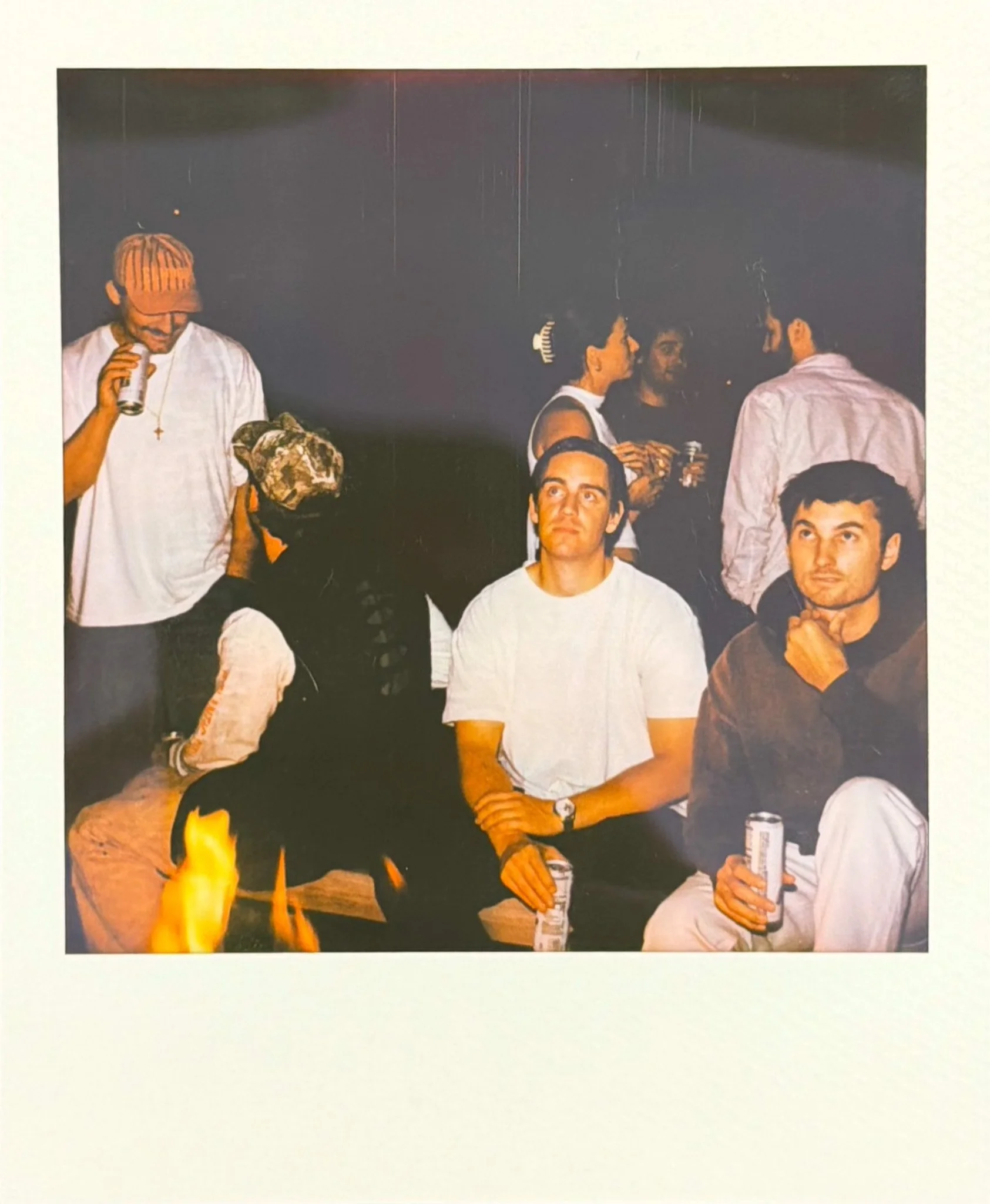 Group of young adults at a party, sitting and standing around a table with drinks, with a fire on the table.