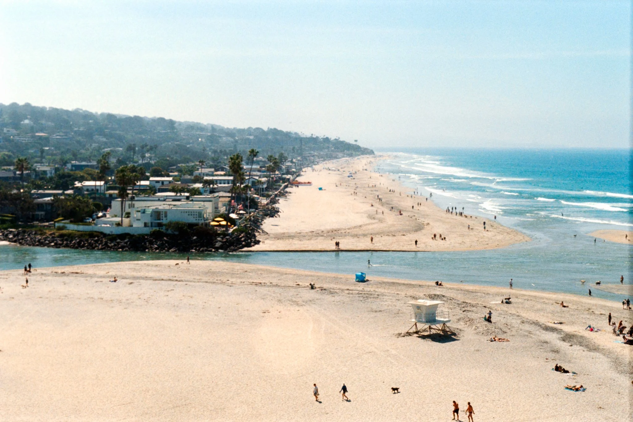 A beach scene with people walking, sitting, and lying on the sandy shore, with houses and palm trees along the coastline and the ocean waves in the background.