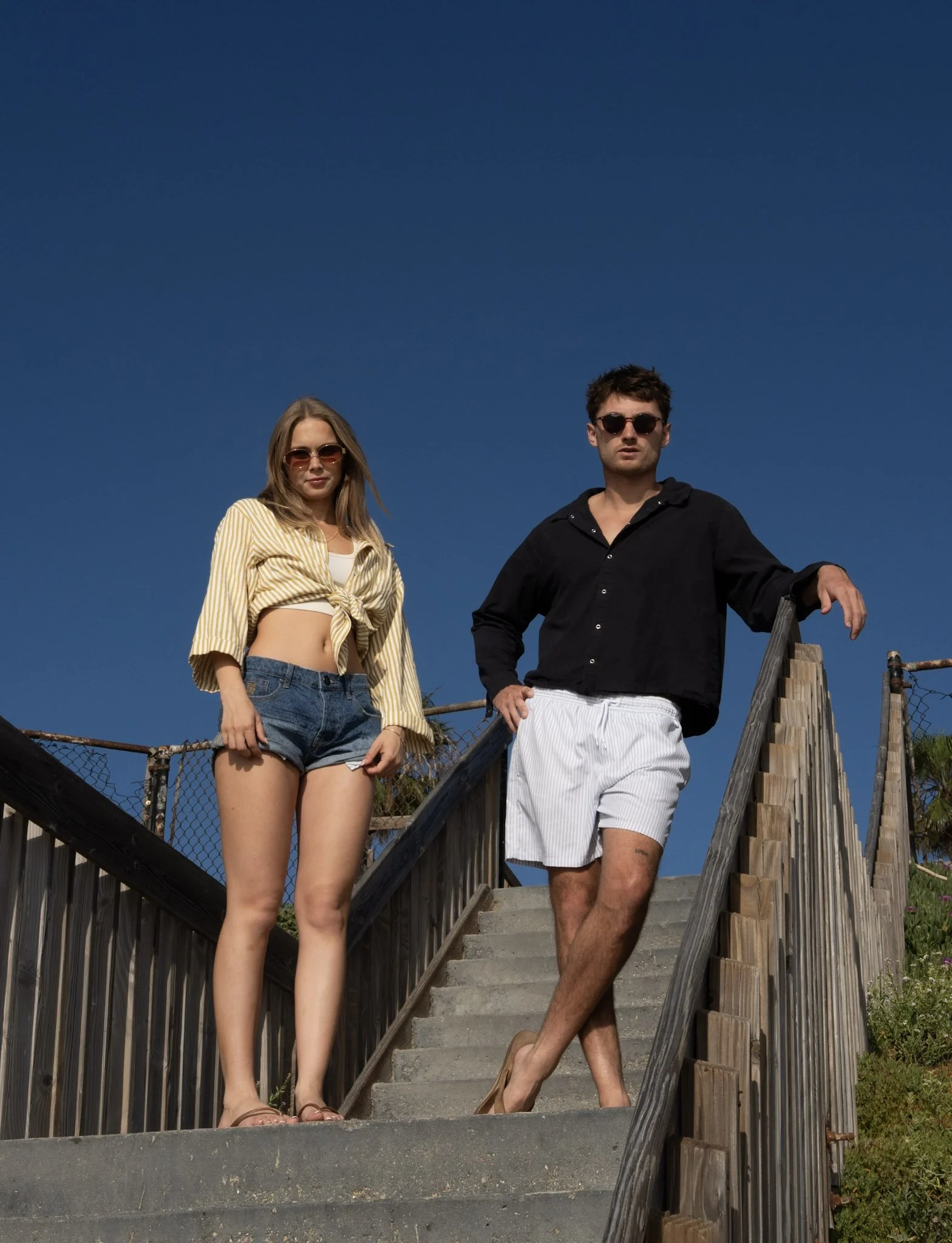 A woman and a man standing on a concrete staircase outdoors under a clear blue sky, both wearing sunglasses, with the woman dressed in a striped yellow shirt tied at the waist and denim shorts, and the man in a black shirt and white striped shorts.