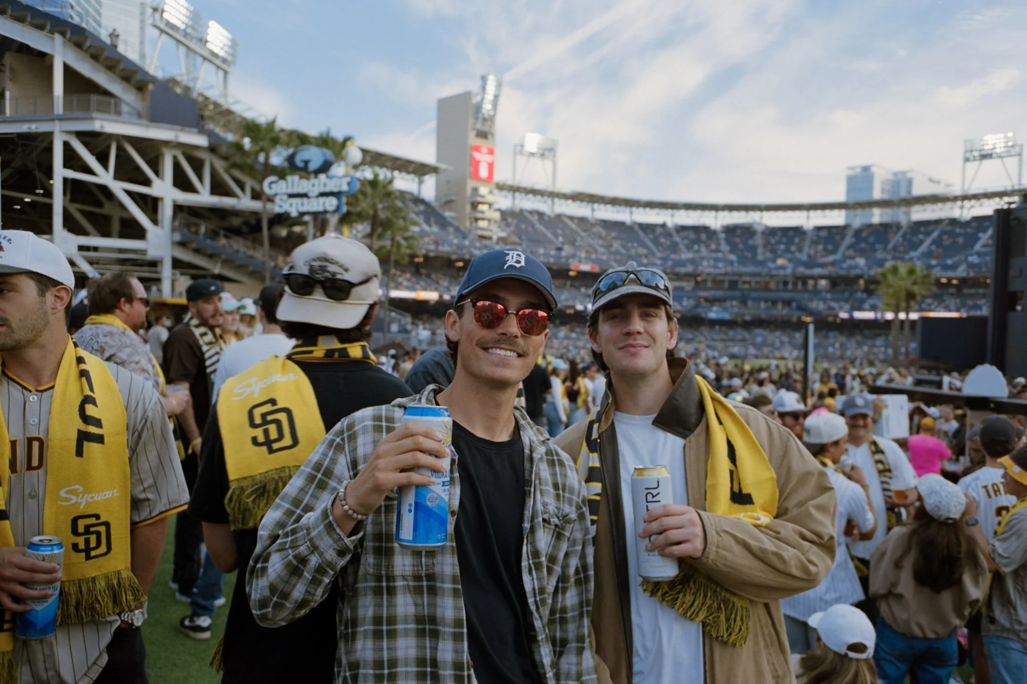 Two young men smiling at a sports stadium with a crowd in the background. They are holding canned beverages, and are wearing casual clothes and sunglasses. The stadium has a sign for Gallagher Square.