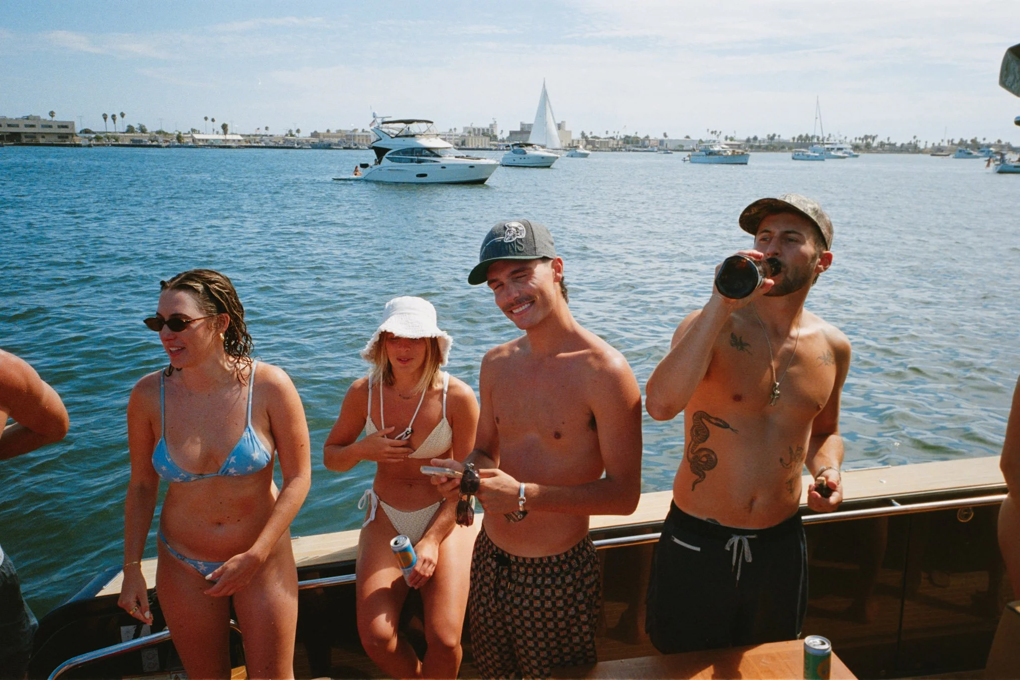 Group of young adults enjoying a boat party in a harbor with boats in the background. Some are wearing swimsuits and hats, and one is drinking from a bottle.